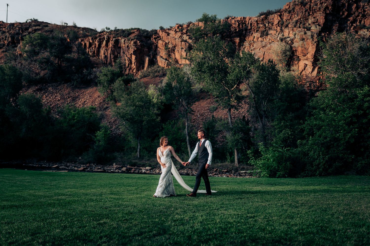 A couple walks hand in hand on a lush green lawn with dramatic red cliffs and trees in the background.