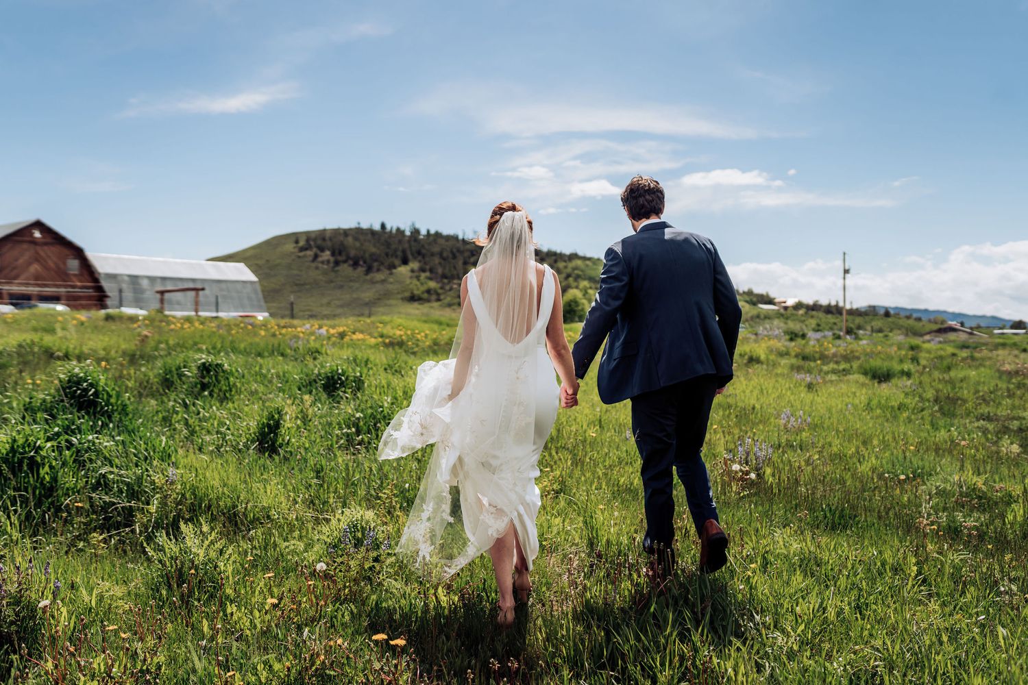 Newlyweds walk hand in hand through a green mountain meadow with a rustic barn in the distance.