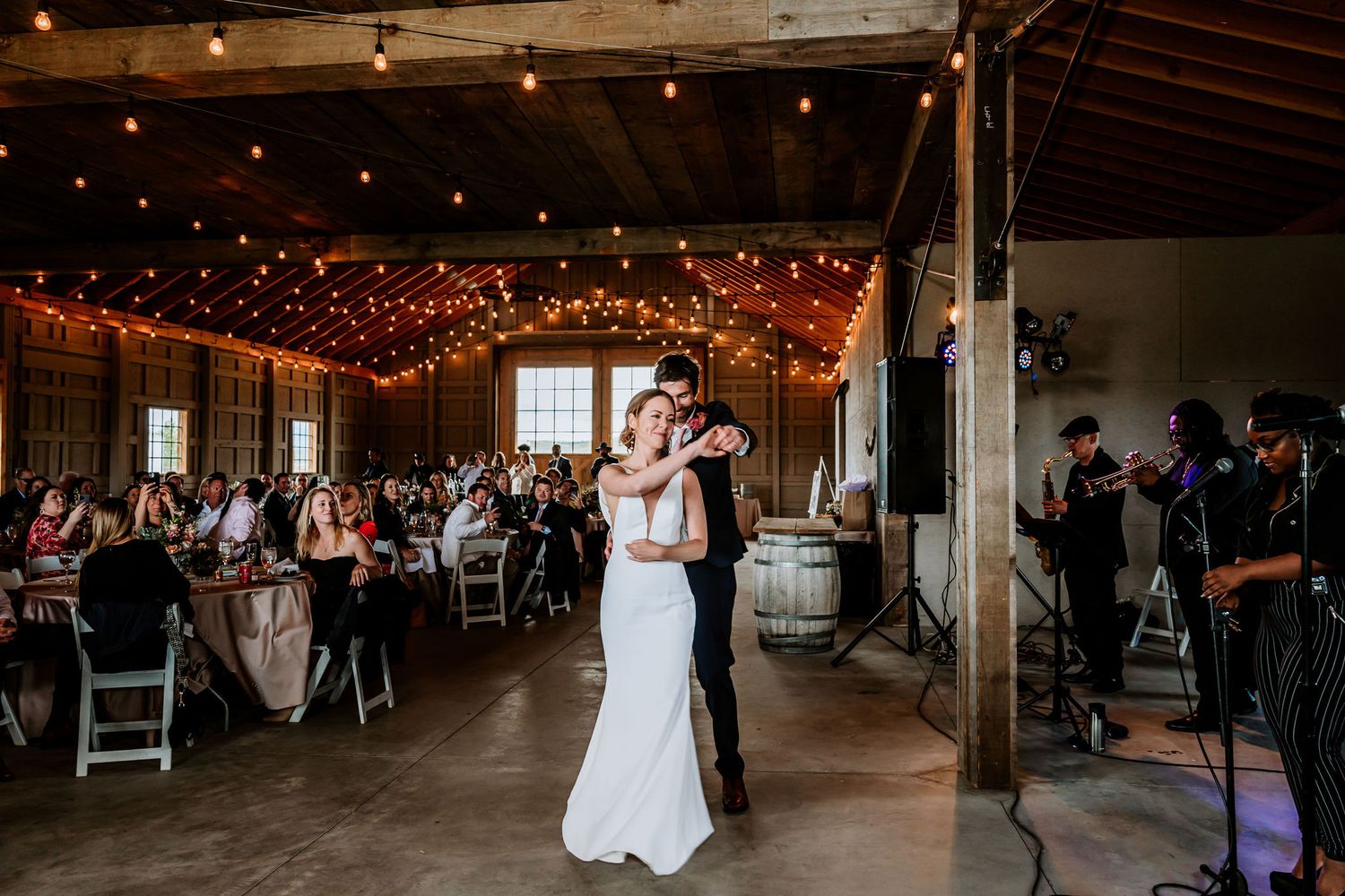 Couple sharing first dance in rustic barn venue with string lights and wedding guests.