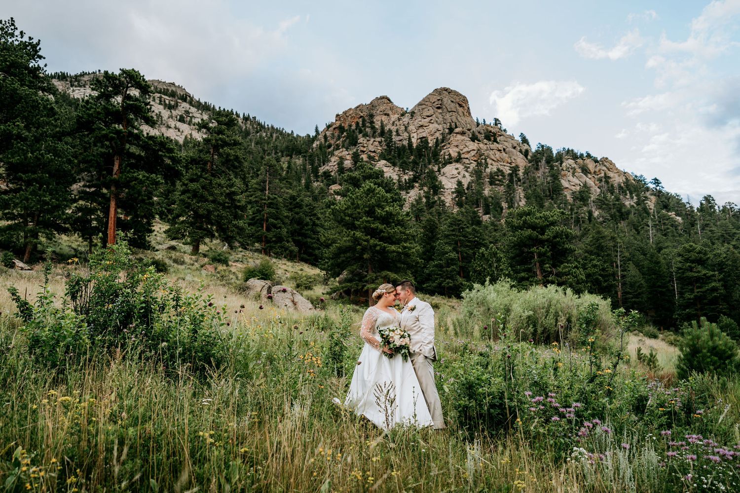 A couple shares an intimate moment in a wildflower meadow with dramatic mountain peaks behind them.