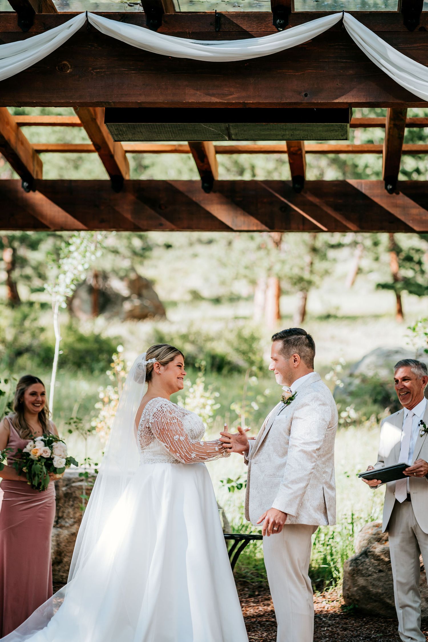 Outdoor wedding ceremony under a wooden pergola decorated with white fabric draping.