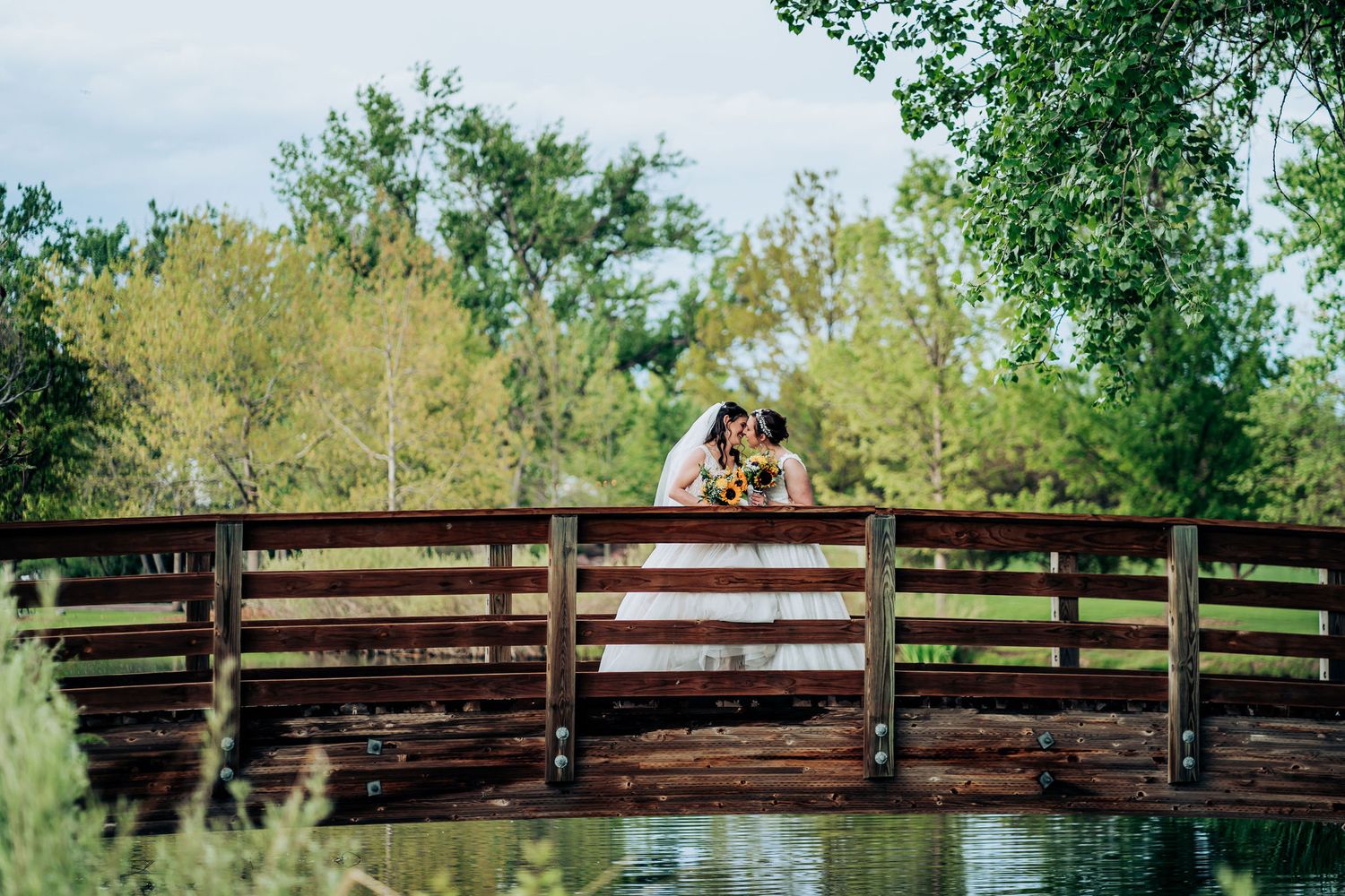 Romantic couple stands on wooden bridge surrounded by lush green trees and nature.
