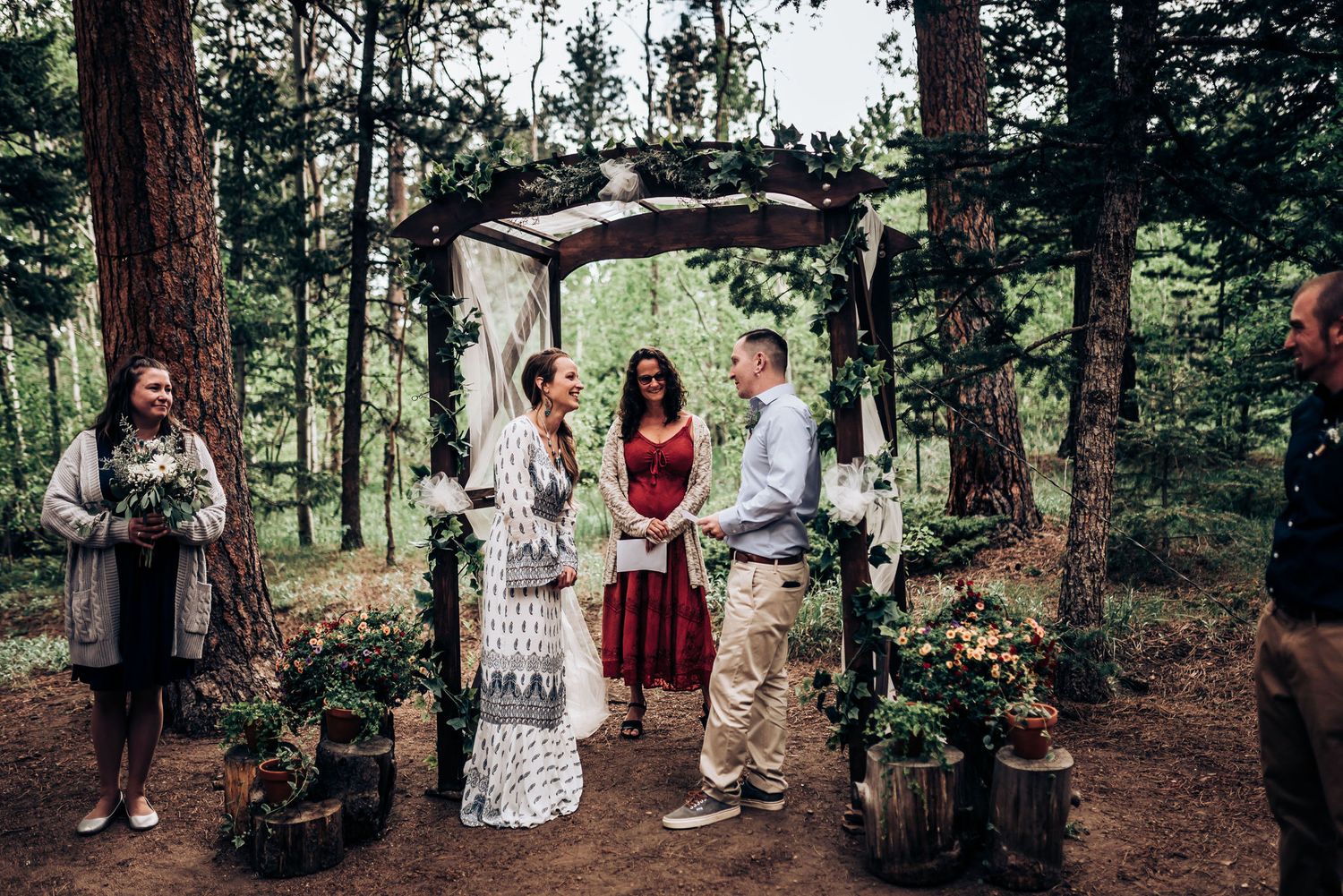 An intimate wedding ceremony takes place under a rustic arch in a pine forest setting.