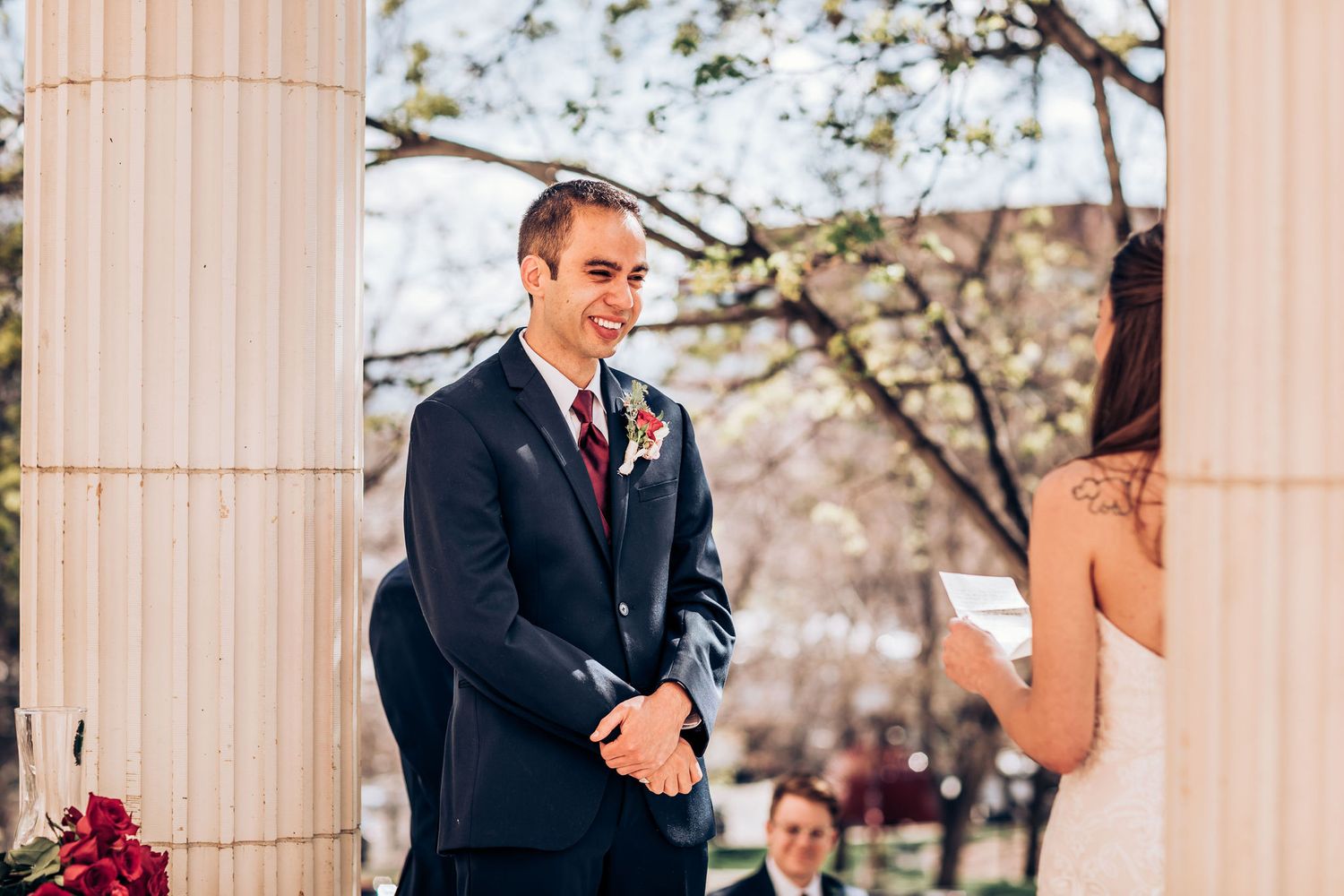 Formal wedding ceremony participant standing between classical columns with spring blooms.