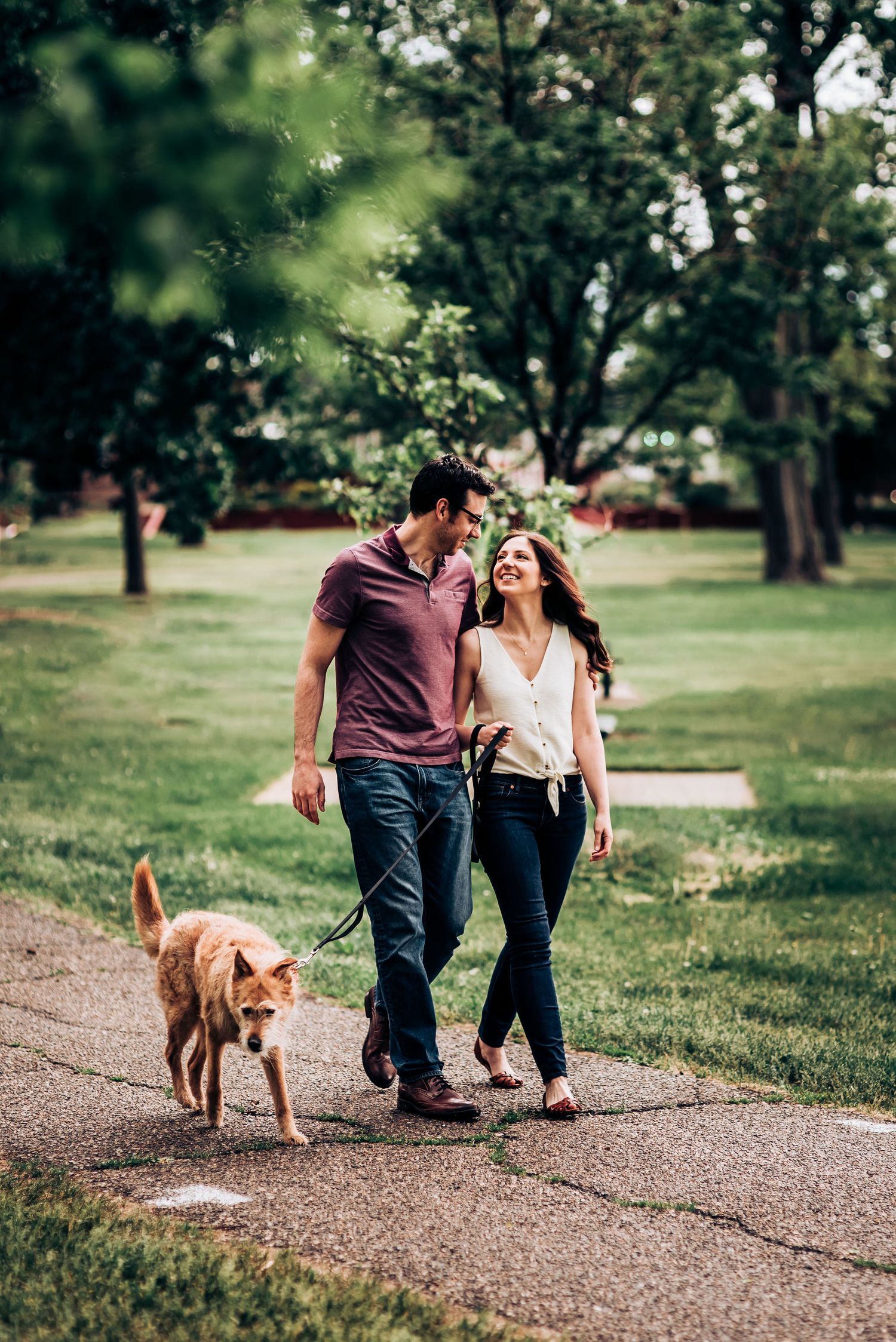A couple walks their golden retriever dog along a tree-lined park path on a sunny day.