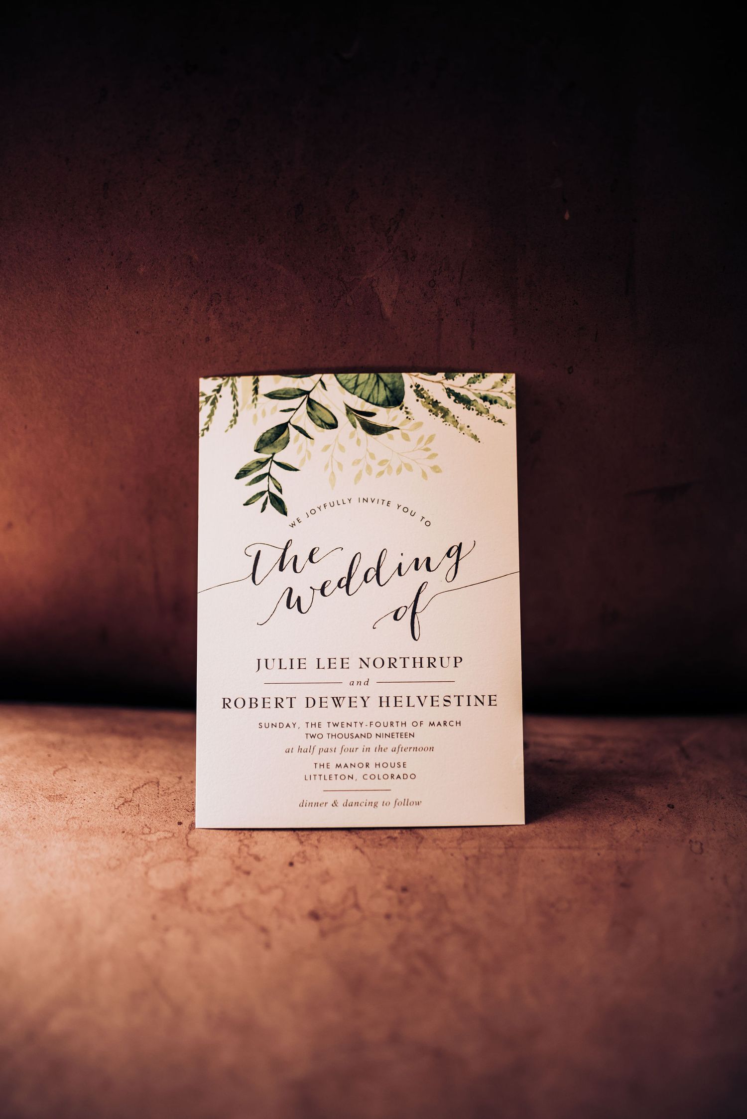 An elegant wedding invitation with greenery design elements on a dark textured background.