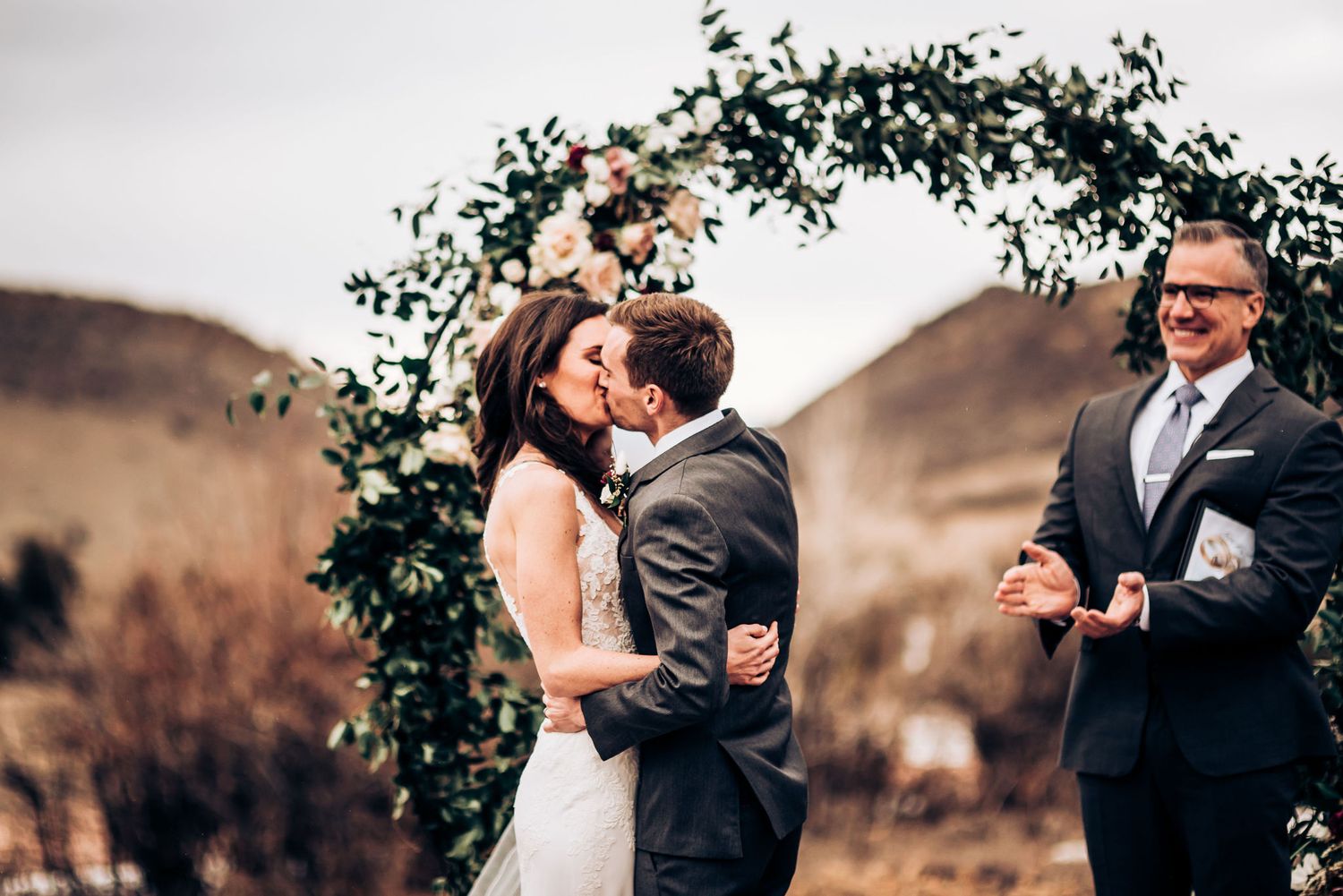 Newlyweds share their first kiss under a leafy arch with mountains in the background.