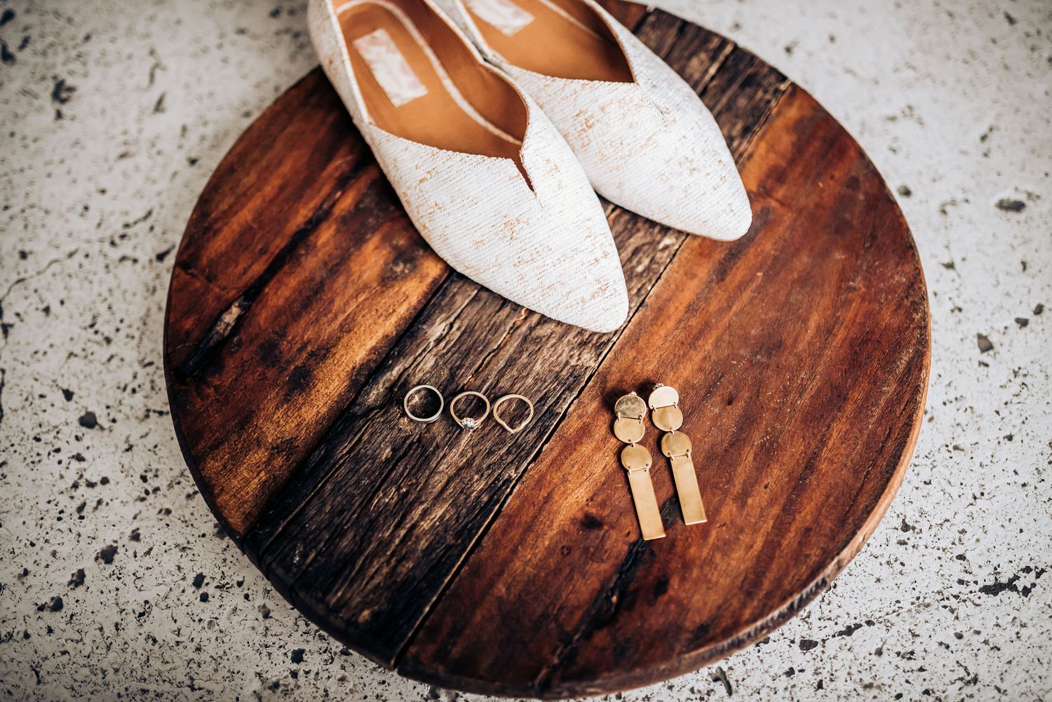 White bridal shoes and wedding rings arranged on a round wooden surface.