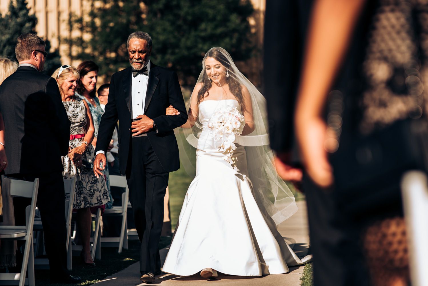 People watching a wedding ceremony outdoors with someone in a white gown walking down the aisle.