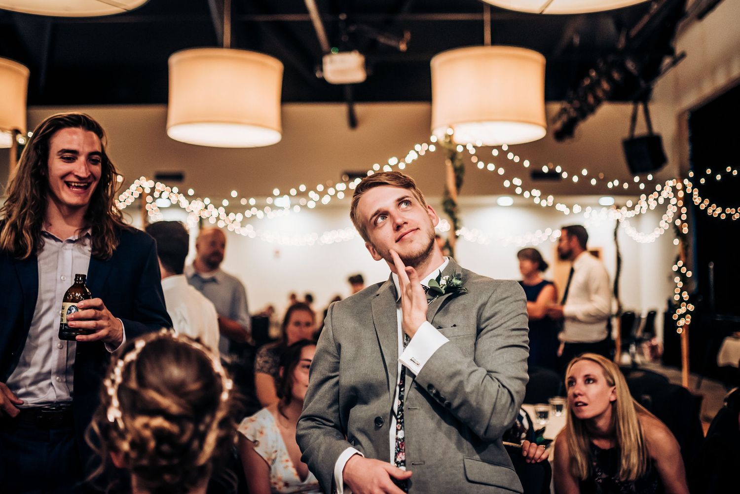 A candid moment during a wedding reception with guests gathered under warm lighting and string lights.