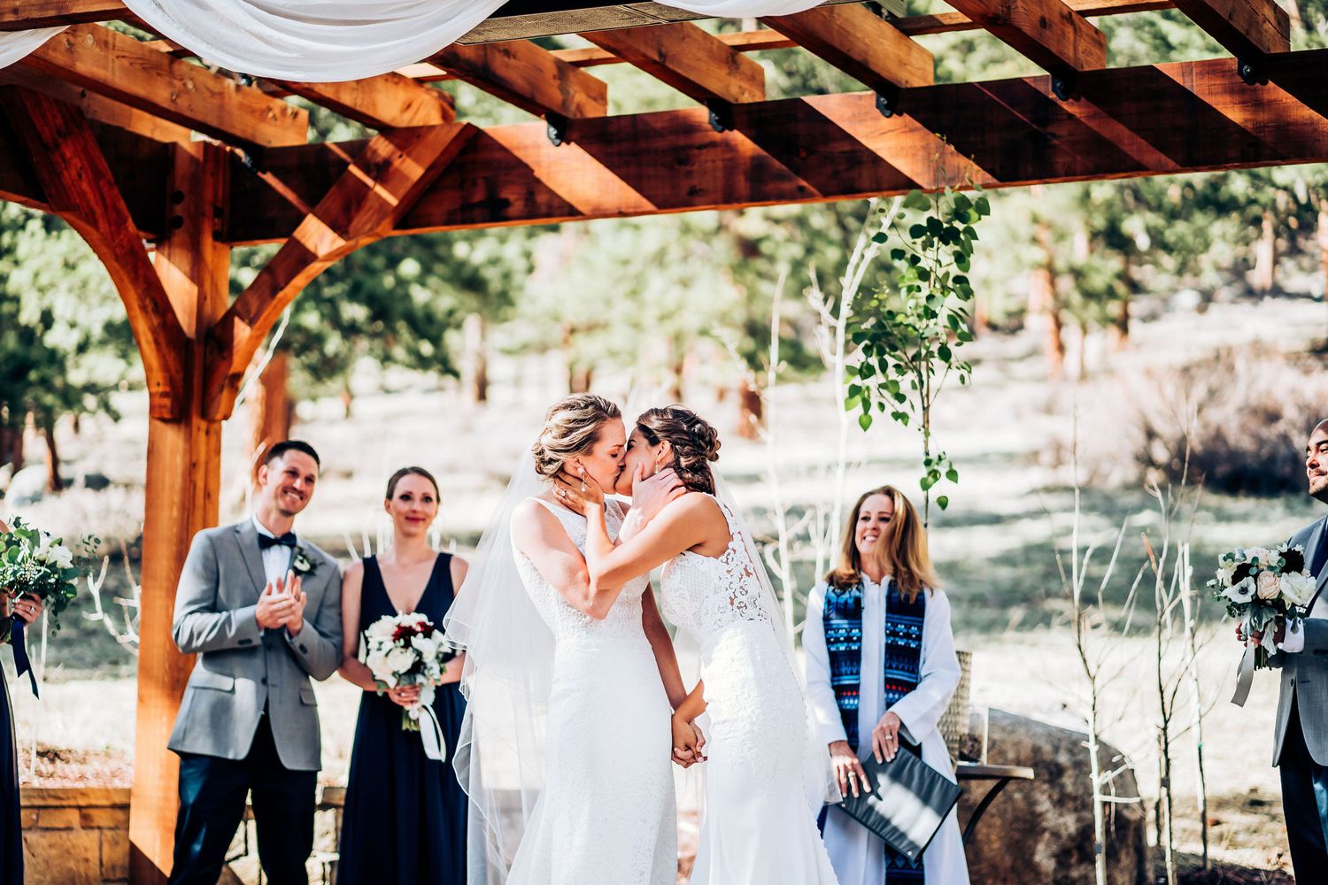 A celebratory moment during an outdoor wedding ceremony under a wooden pergola with guests watching.