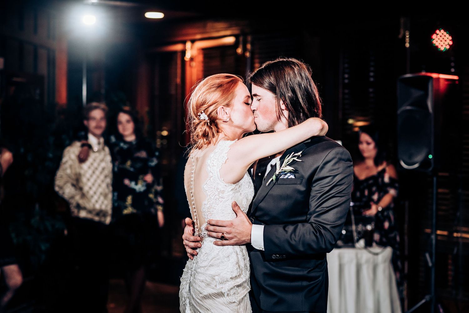 A couple shares an intimate kiss during their wedding dance as guests watch in the background.