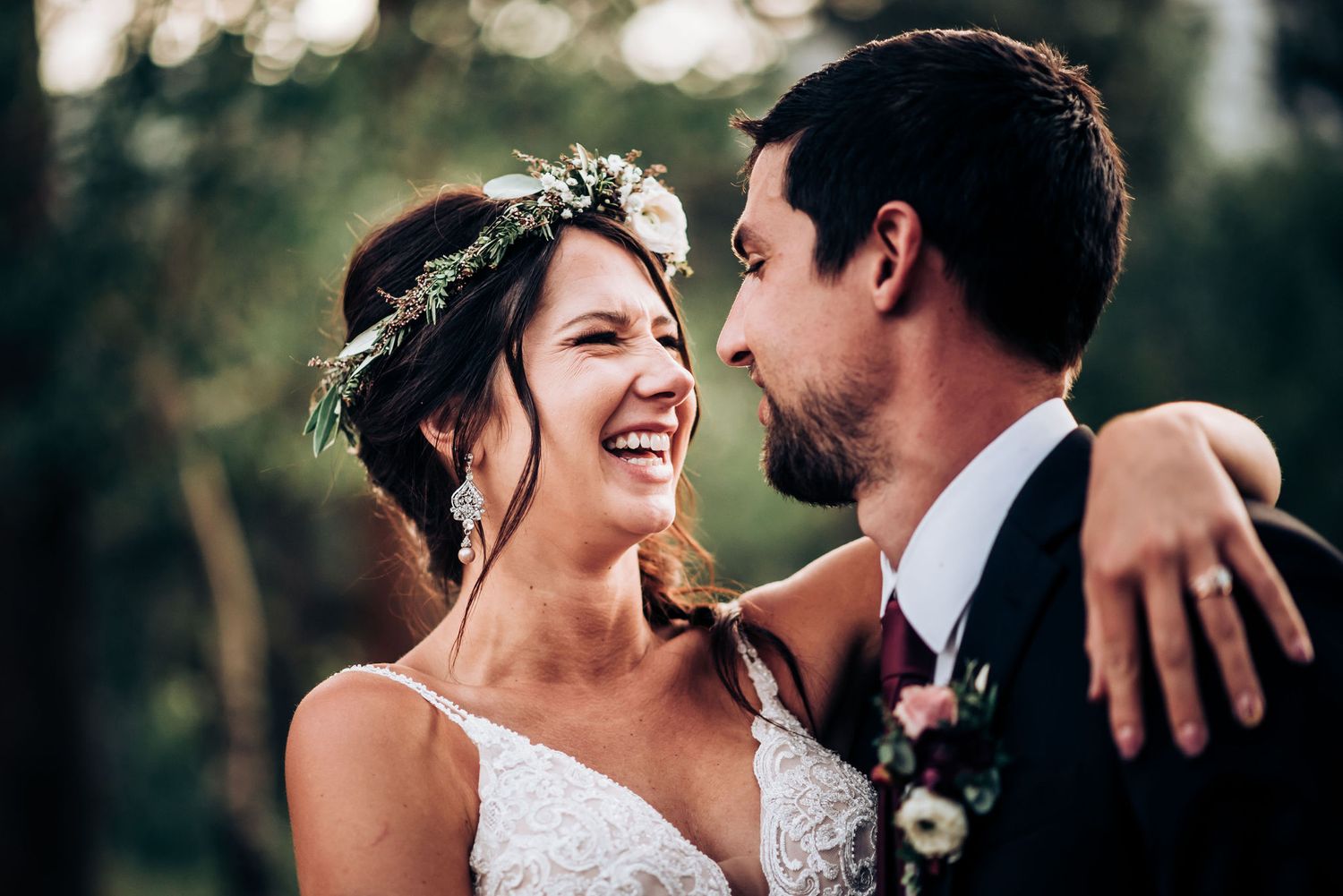Romantic candid moment between newlyweds laughing together outdoors wearing a floral crown and formal attire.