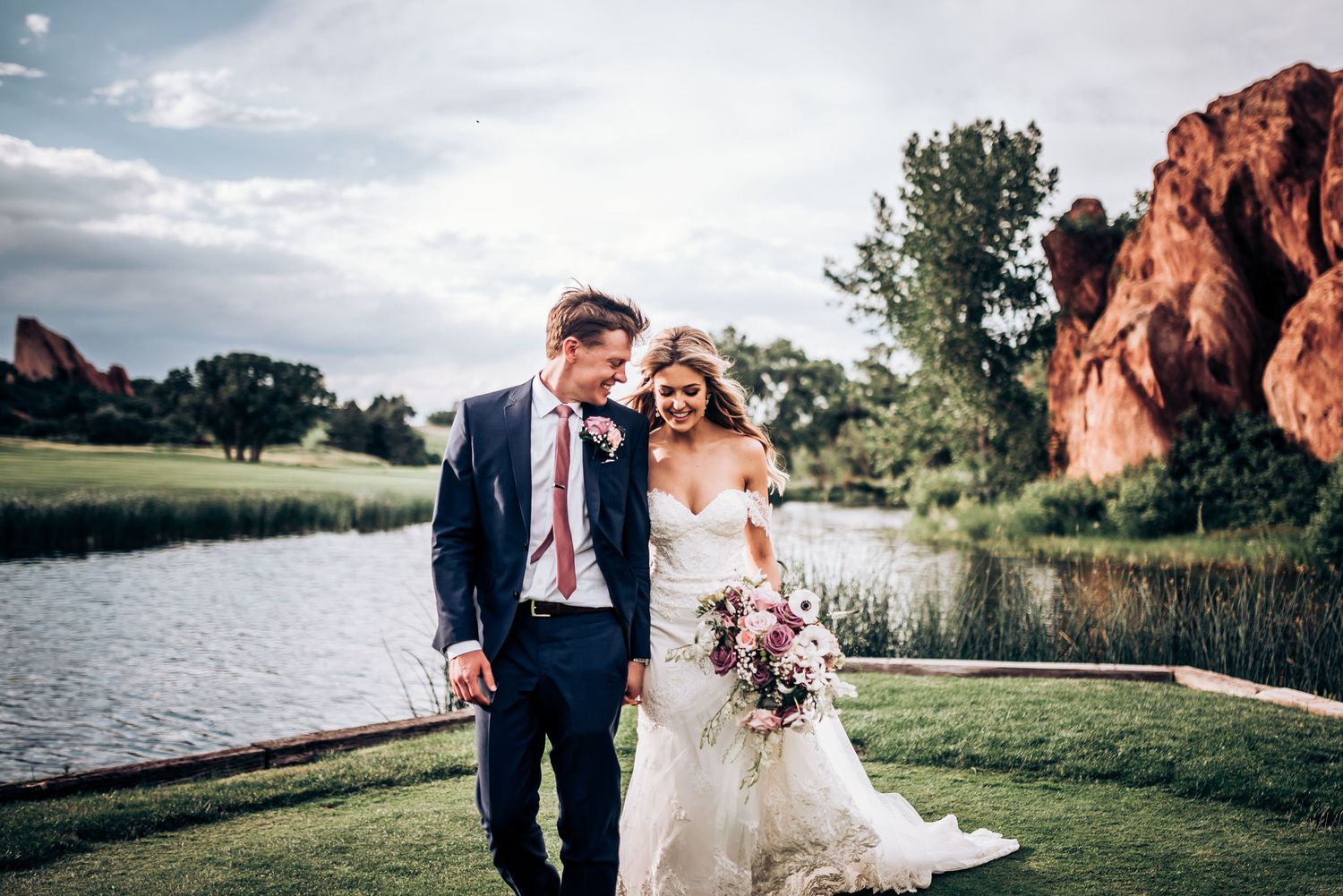 Joyful wedding portrait with vibrant bouquet against red rock formations and lake.