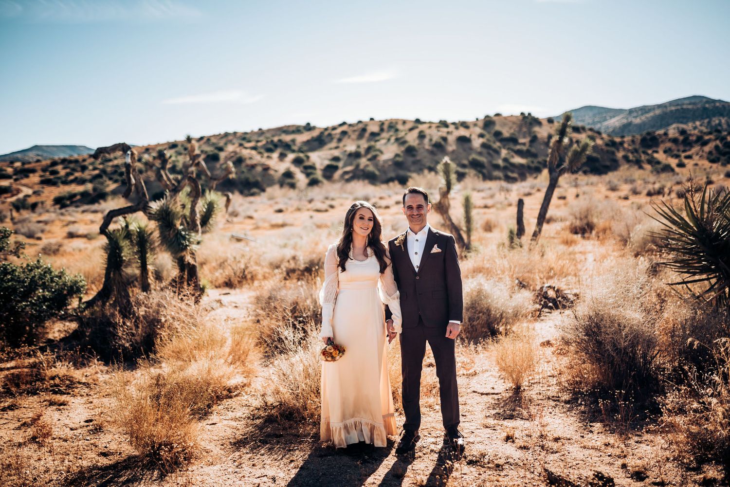 Desert wedding portrait with couple standing among cacti in golden afternoon light.