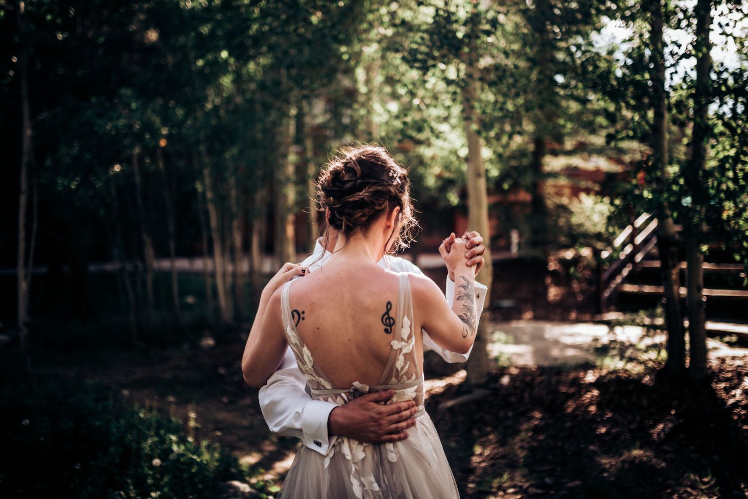 Back view of a bride in a backless lace wedding gown standing in a sunlit forest setting.