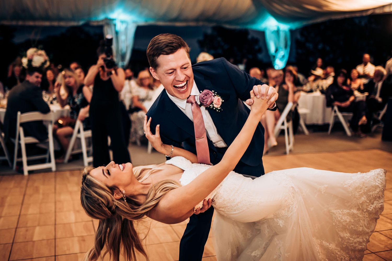 Newlyweds perform a dramatic dip during their wedding dance on a wooden floor under draped fabric.