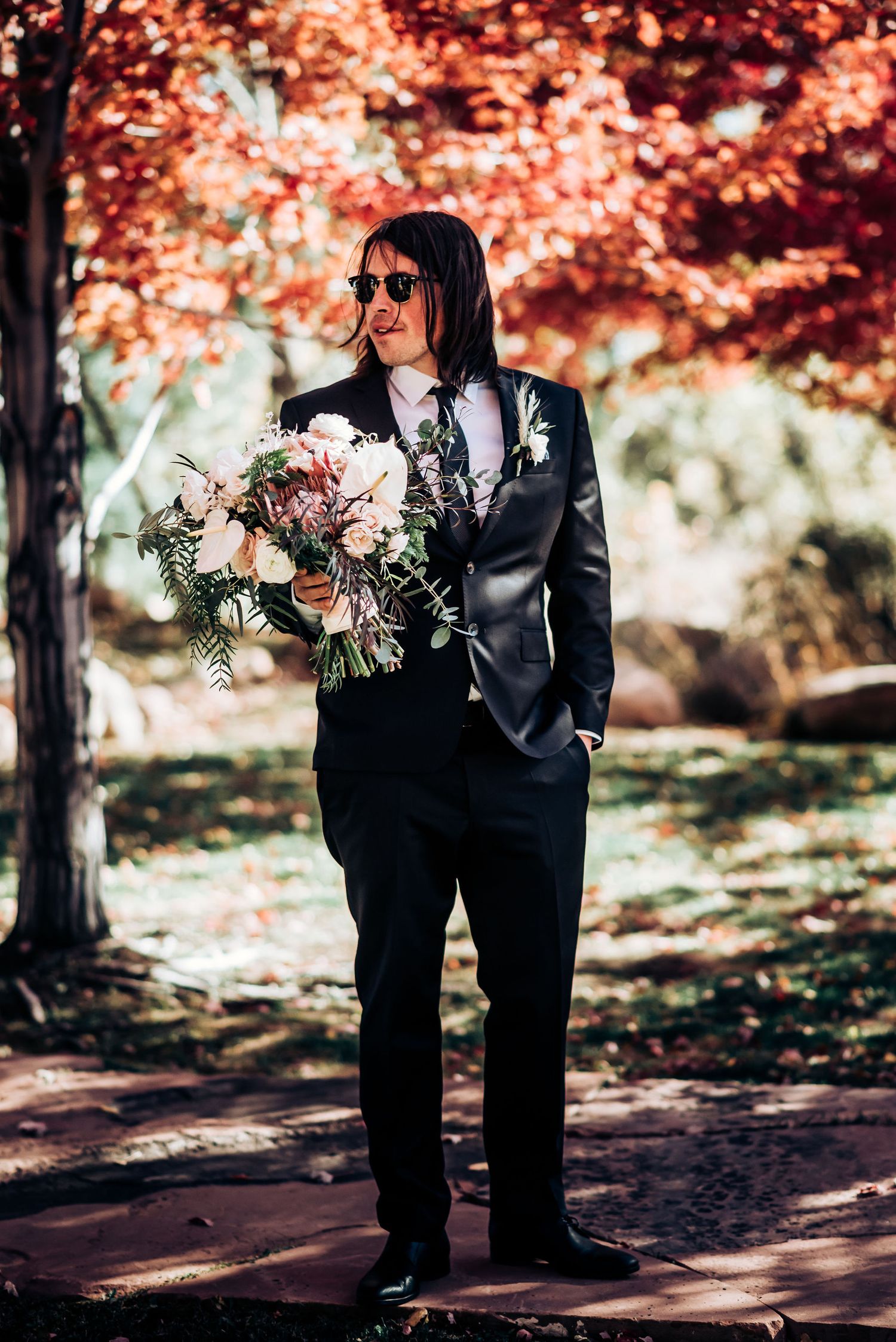 Wedding party member in black suit holds pink and white floral bouquet under red maple trees.