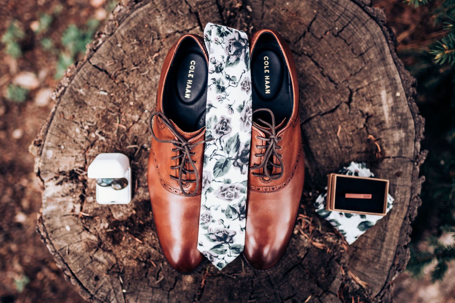 Brown leather dress shoes with floral tie and accessories arranged on wooden surface.
