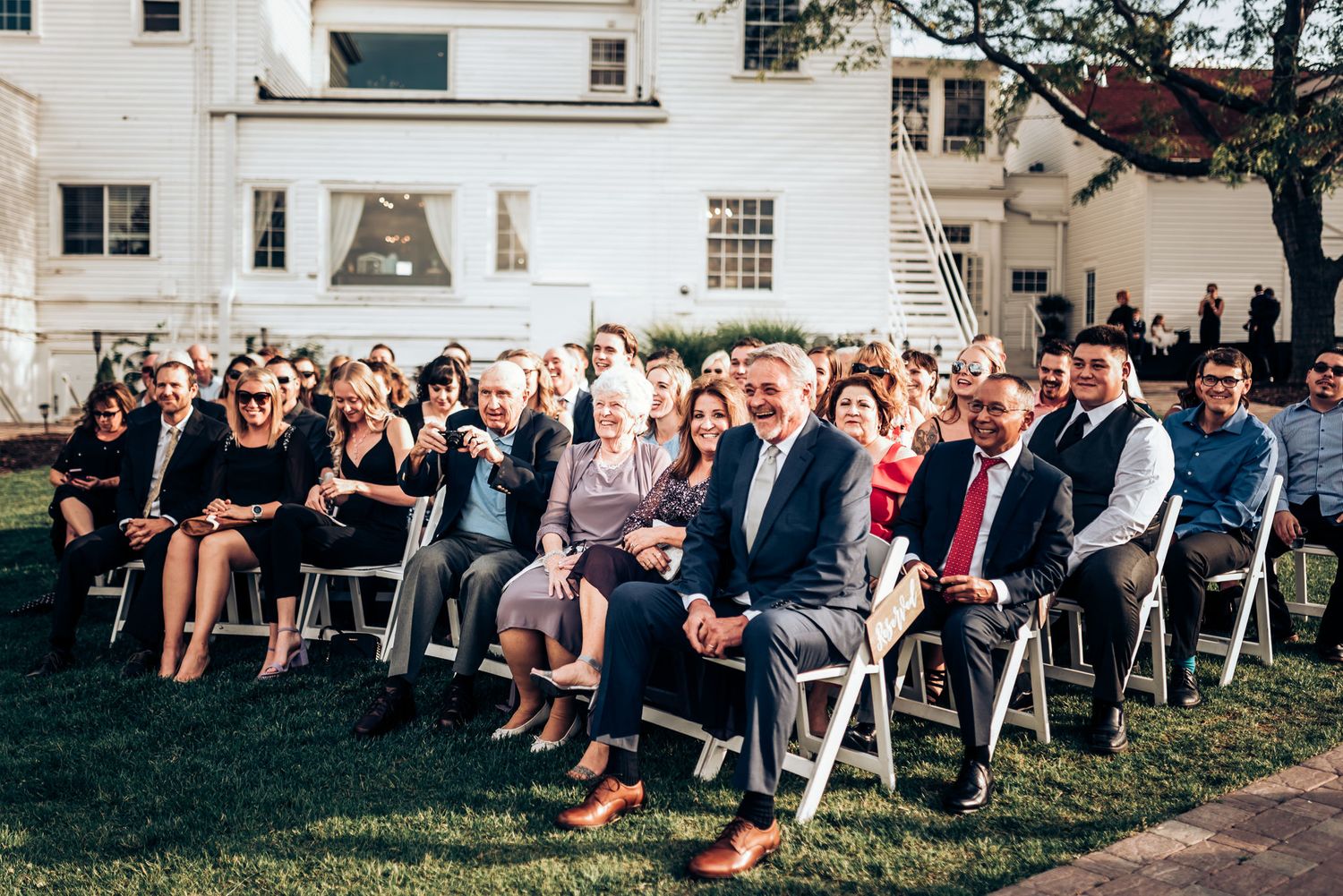 Wedding ceremony guests seated outdoors on white chairs at elegant venue.