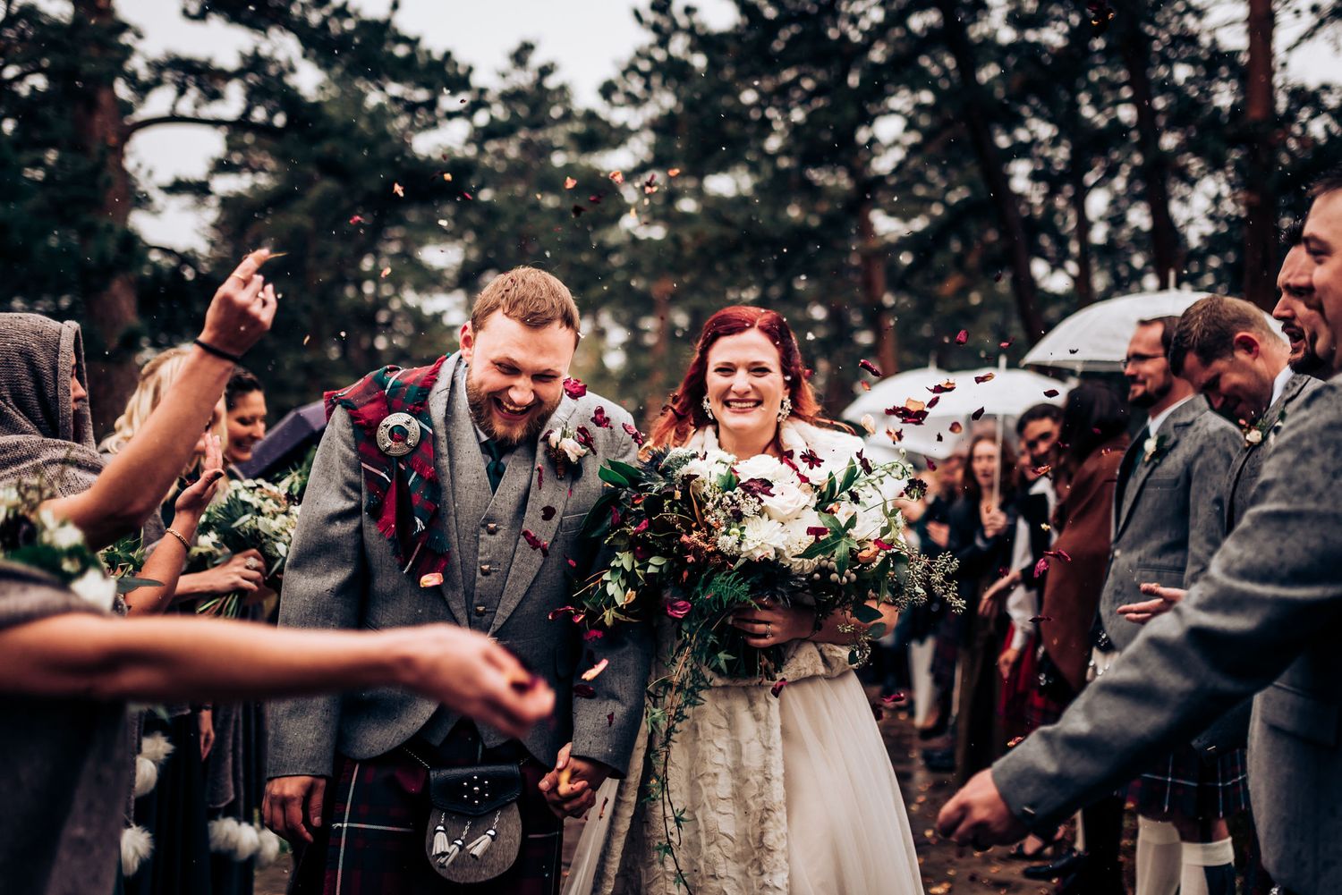 Newlyweds walking through cheering guests with umbrellas in a pine forest setting.