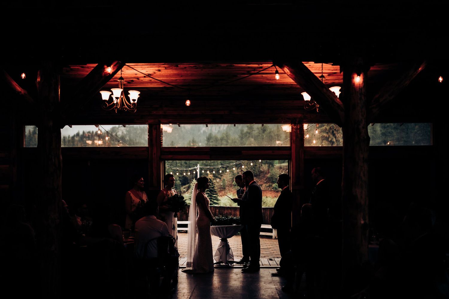 A silhouette of wedding ceremony attendees gathered under a rustic wooden structure at sunset.