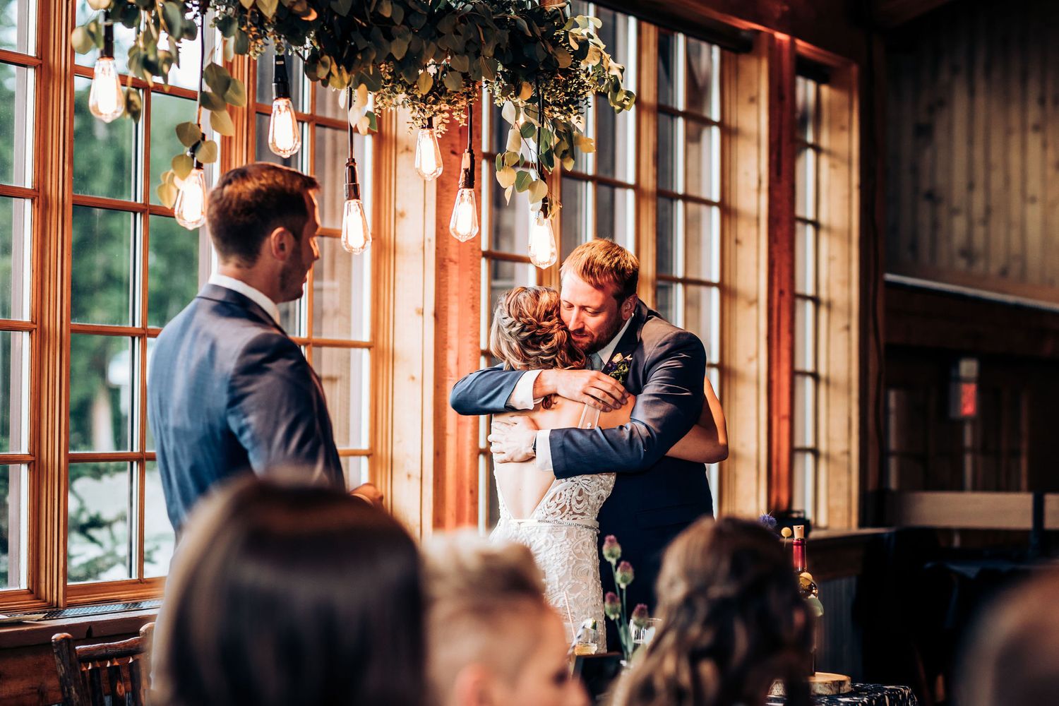 Wedding guests share an emotional toast moment in a rustic venue with large windows.