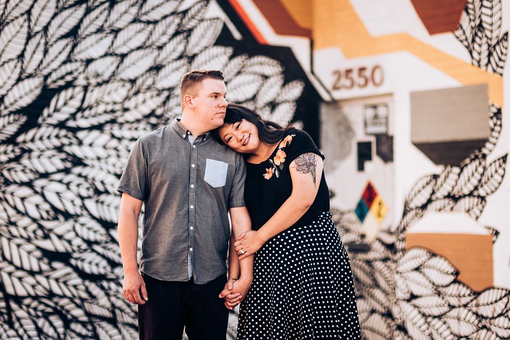 Couple poses playfully against geometric mural wall in urban setting.