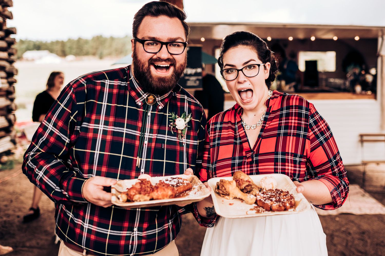 Coordinated pair in matching plaid shirts display plates of food at outdoor gathering.
