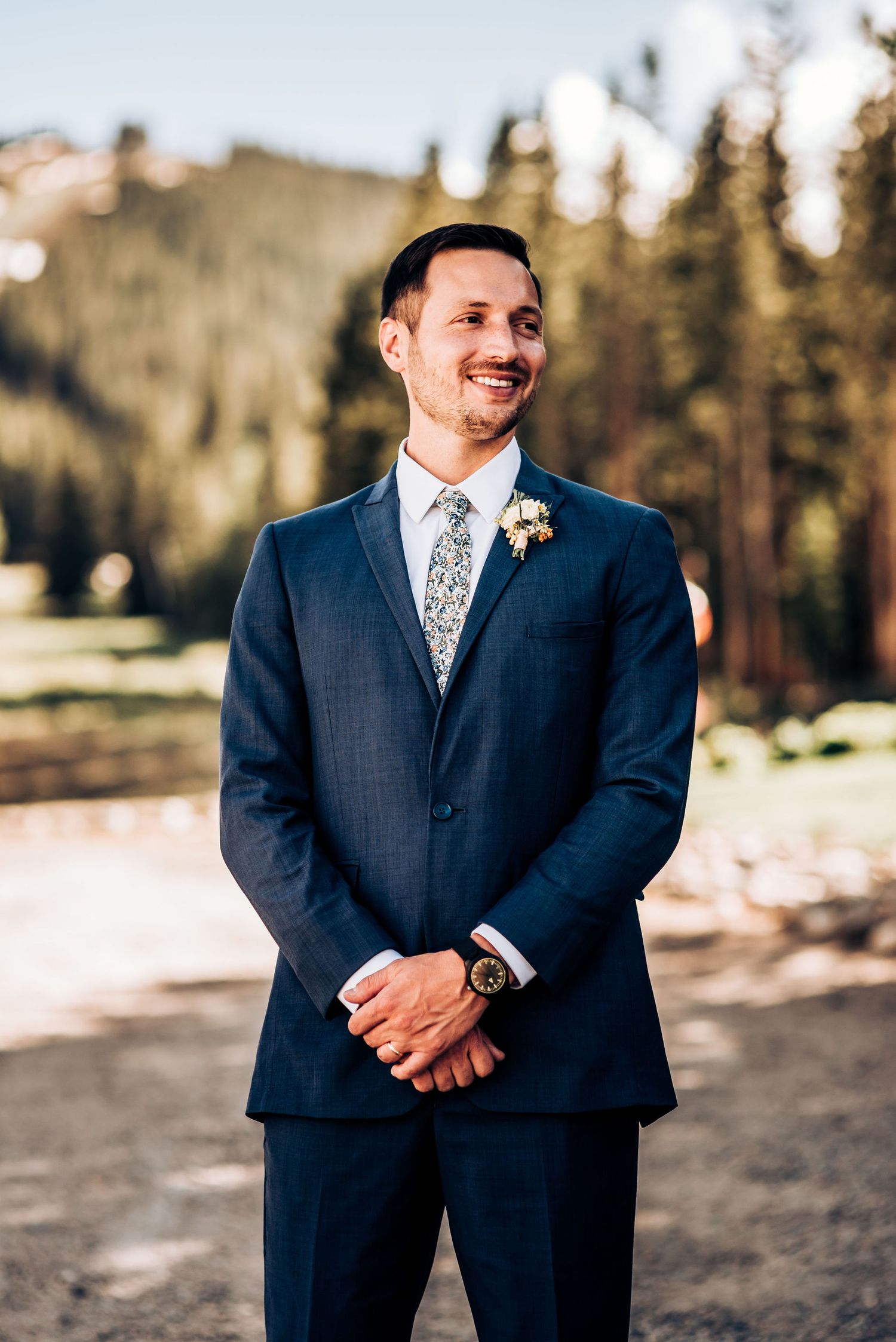 Groom in navy suit with floral boutonniere smiling against mountain backdrop.