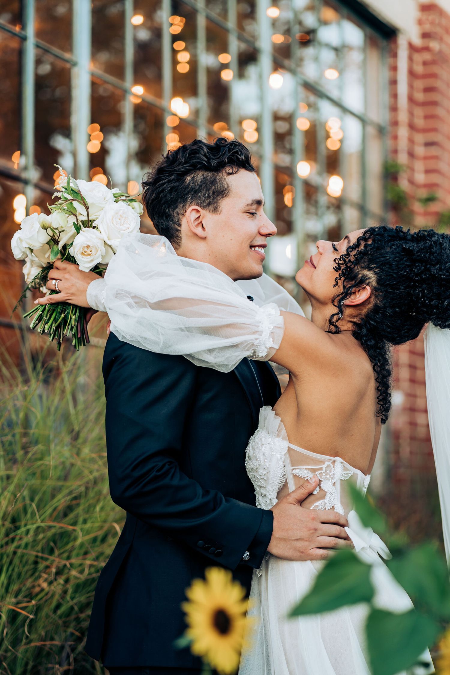 A romantic wedding portrait with sunflowers and string lights decorating the brick building backdrop.