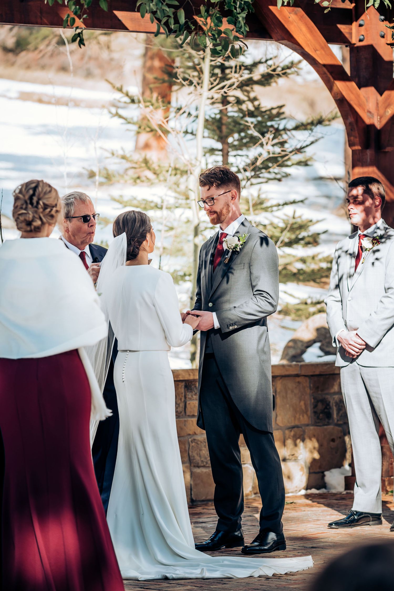 Intimate mountain wedding ceremony under rustic wooden arch with snow in background.