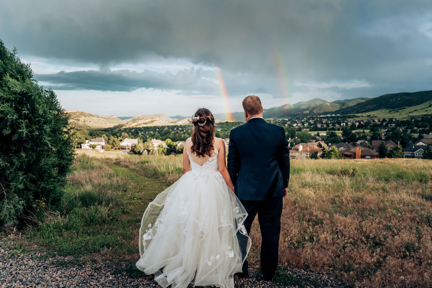 A wedding couple gazes at a stunning rainbow while standing on a hilltop overlooking a valley.