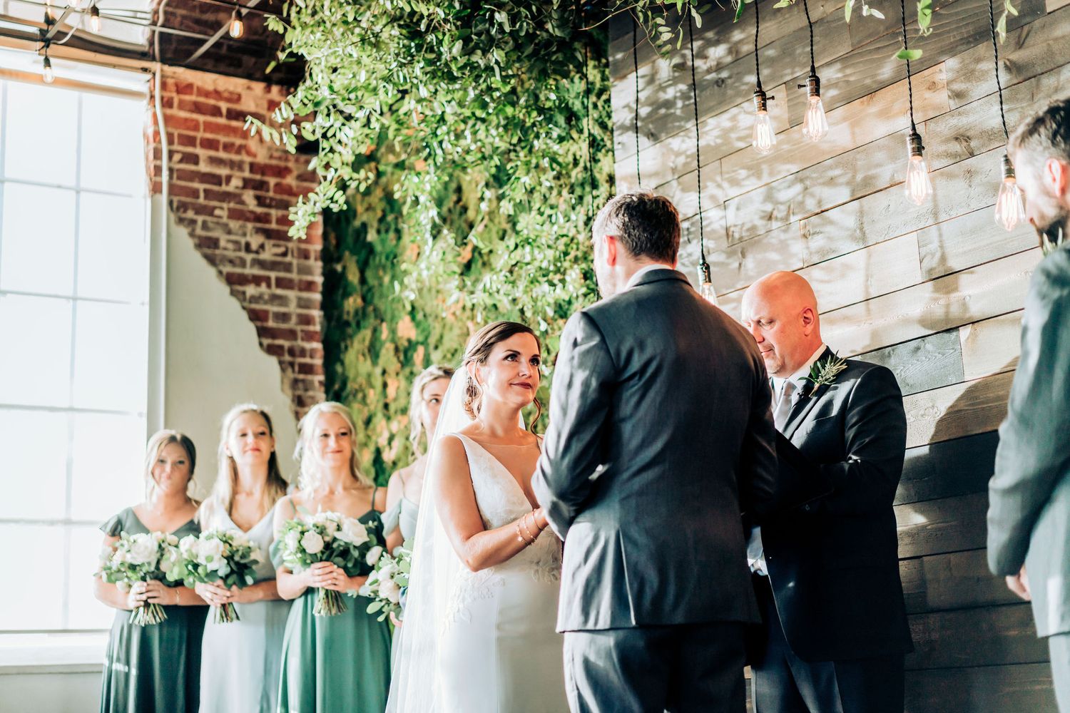 Indoor wedding ceremony with brick walls, hanging greenery and attendants in sage green dresses.