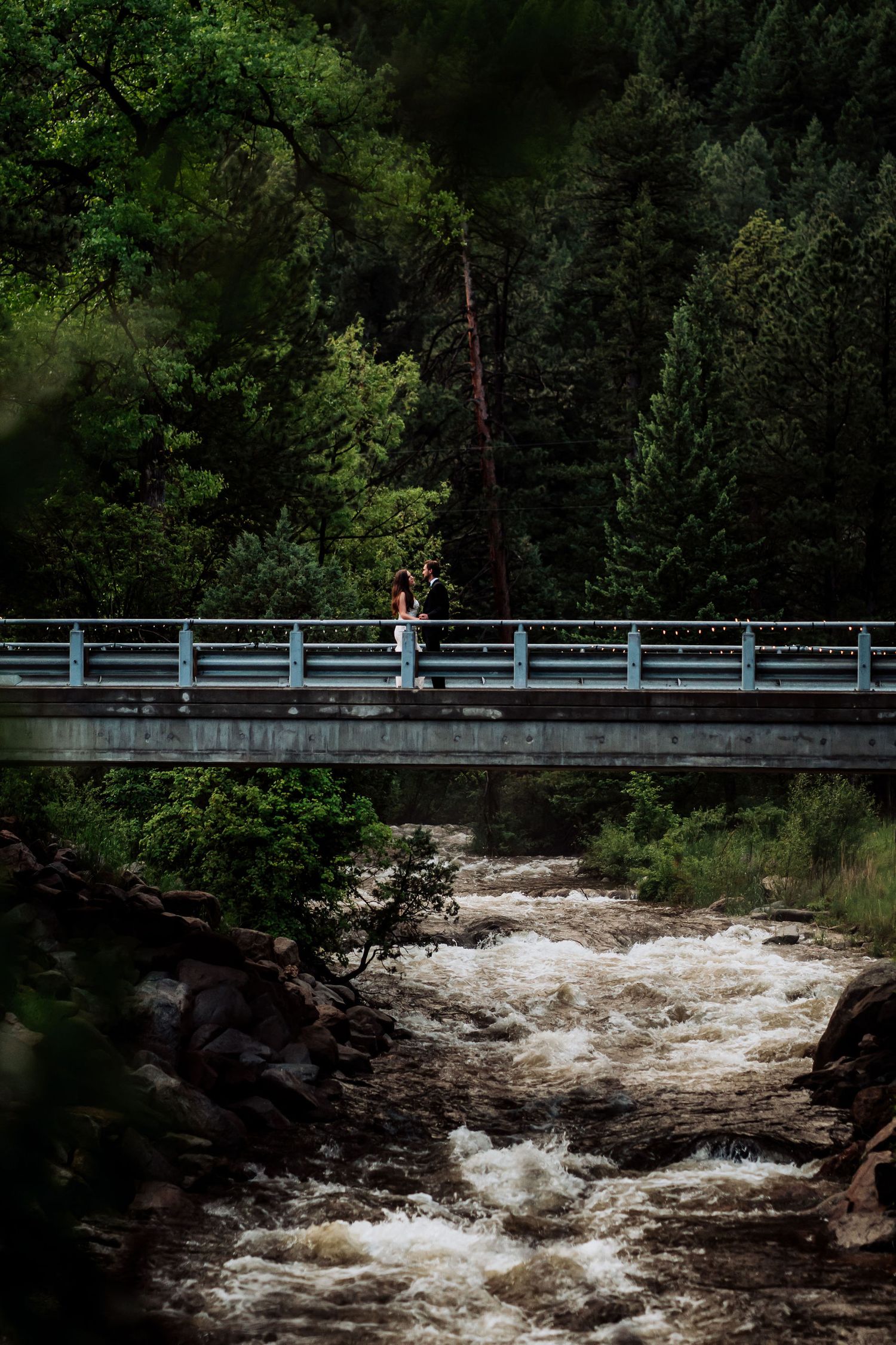 Couple stands on bridge over rushing river surrounded by lush forest.