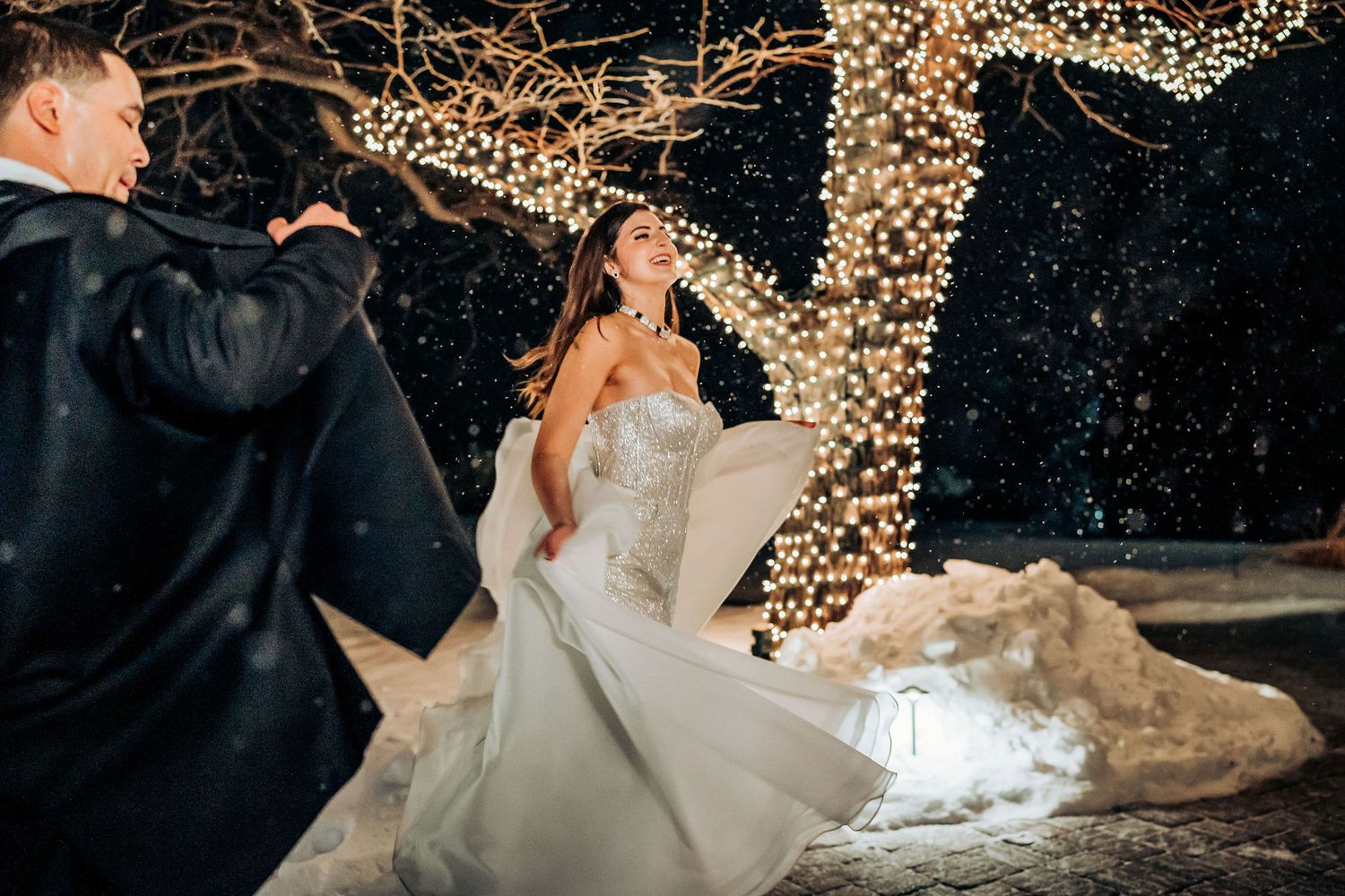 Magical winter wedding portrait with couple dancing under illuminated trees in falling snow.