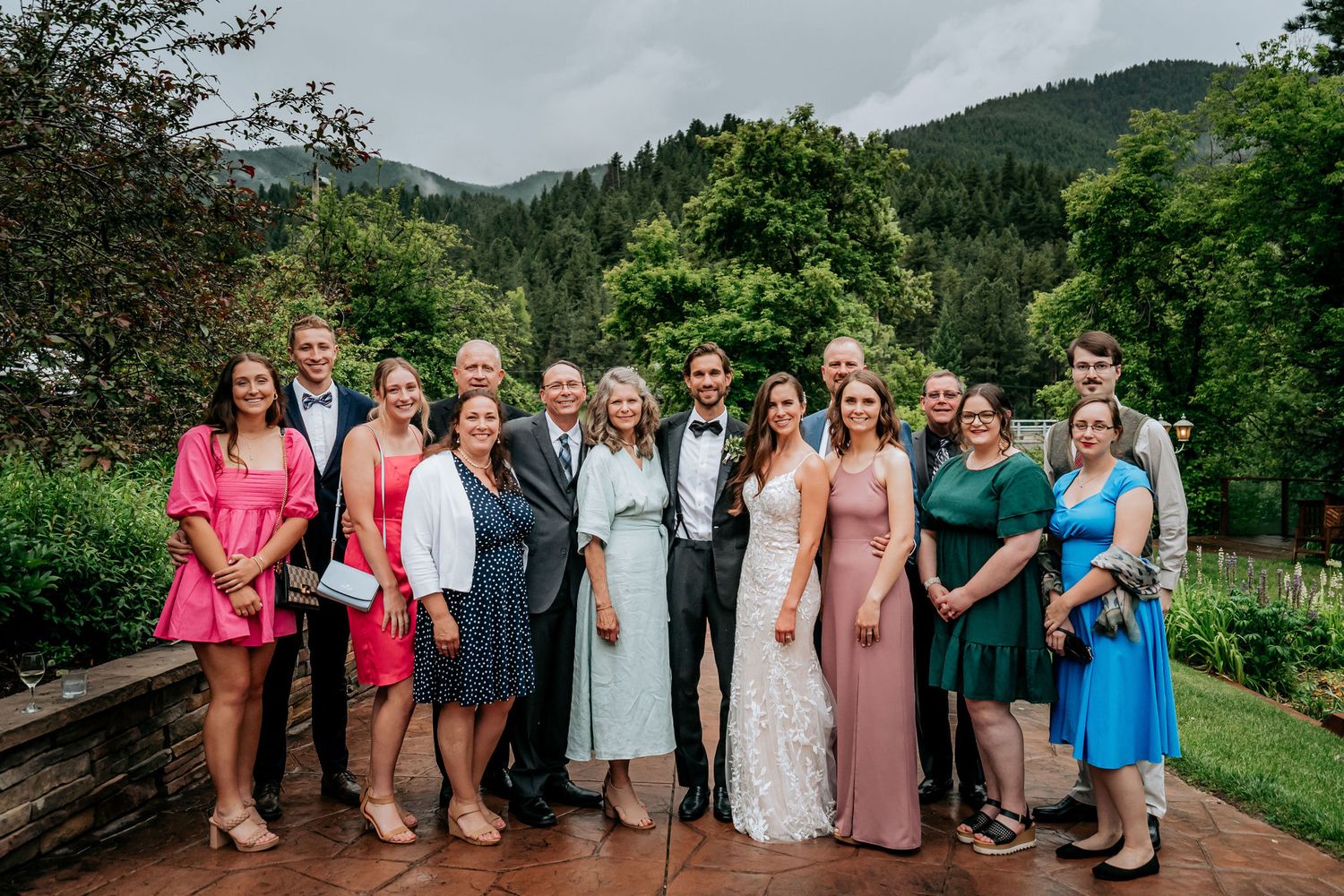 Wedding party poses for group photo on mountain deck with scenic forest backdrop.