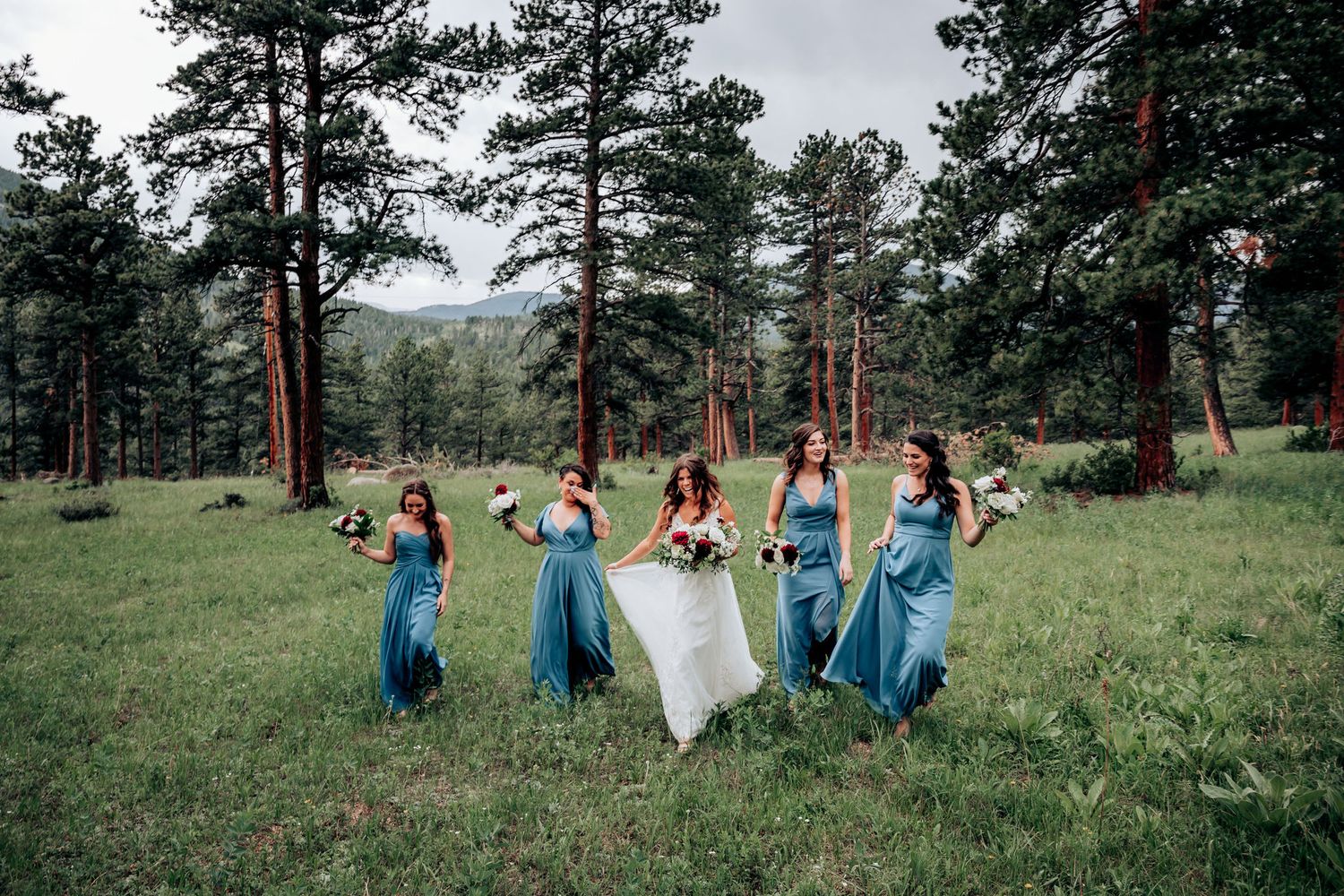 Bridesmaids in blue dresses dance joyfully with bride in mountain forest clearing.