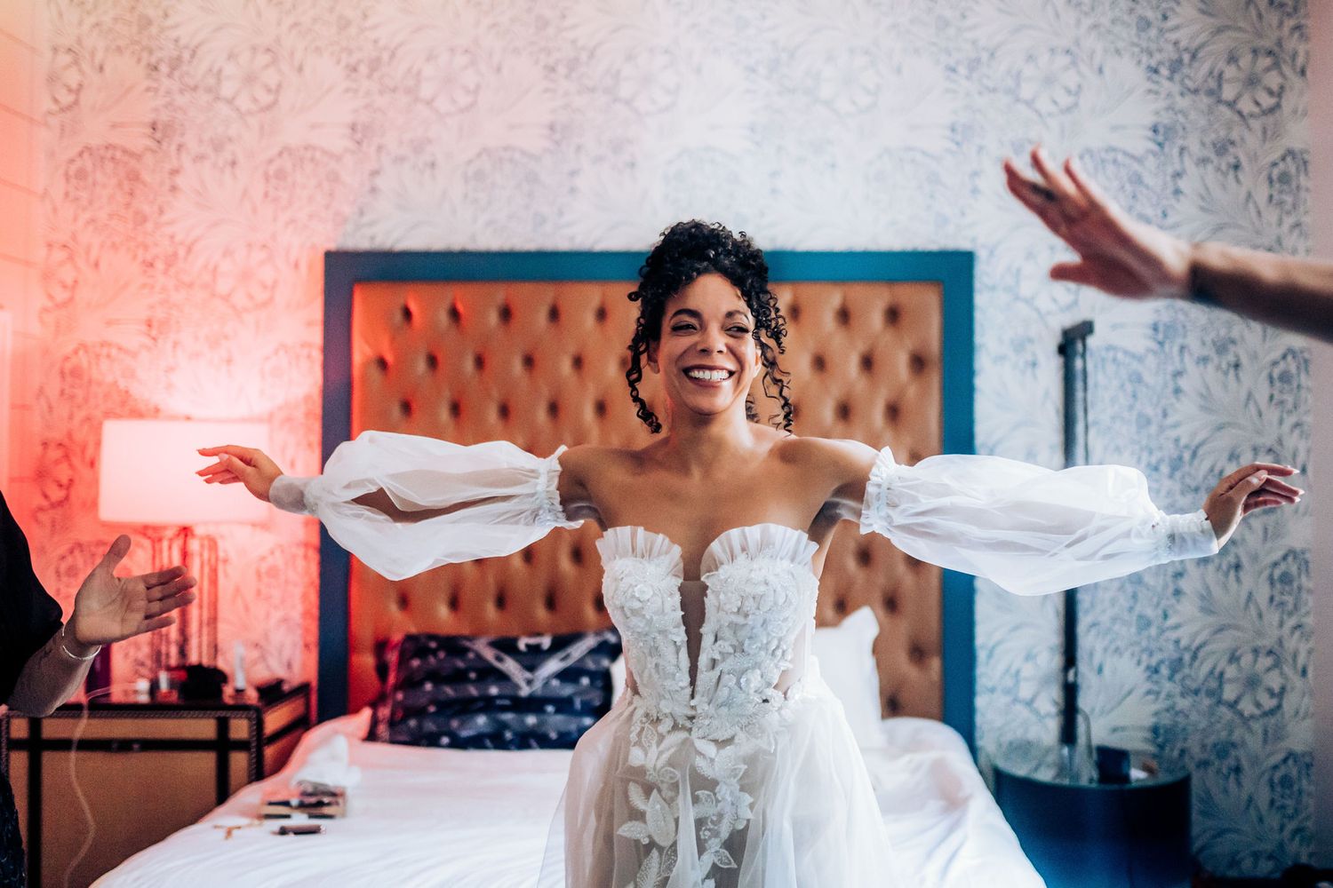 A bride in a white strapless gown spreads her arms wide with joy in a decorated room.