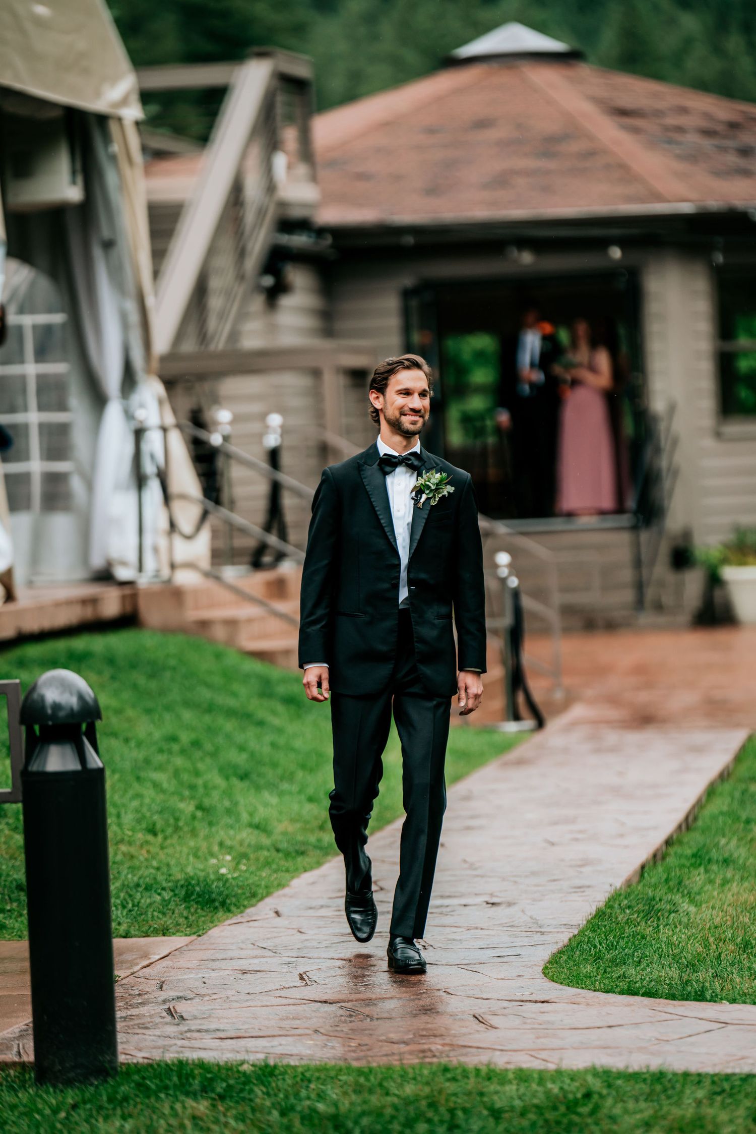 A formal wedding attendant walks along a stone path outside a rustic venue building.