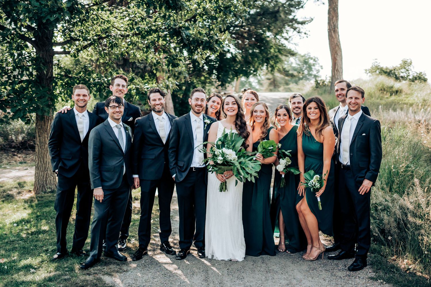 Full wedding party poses together outdoors with bridesmaids in dark green and groomsmen in black suits.