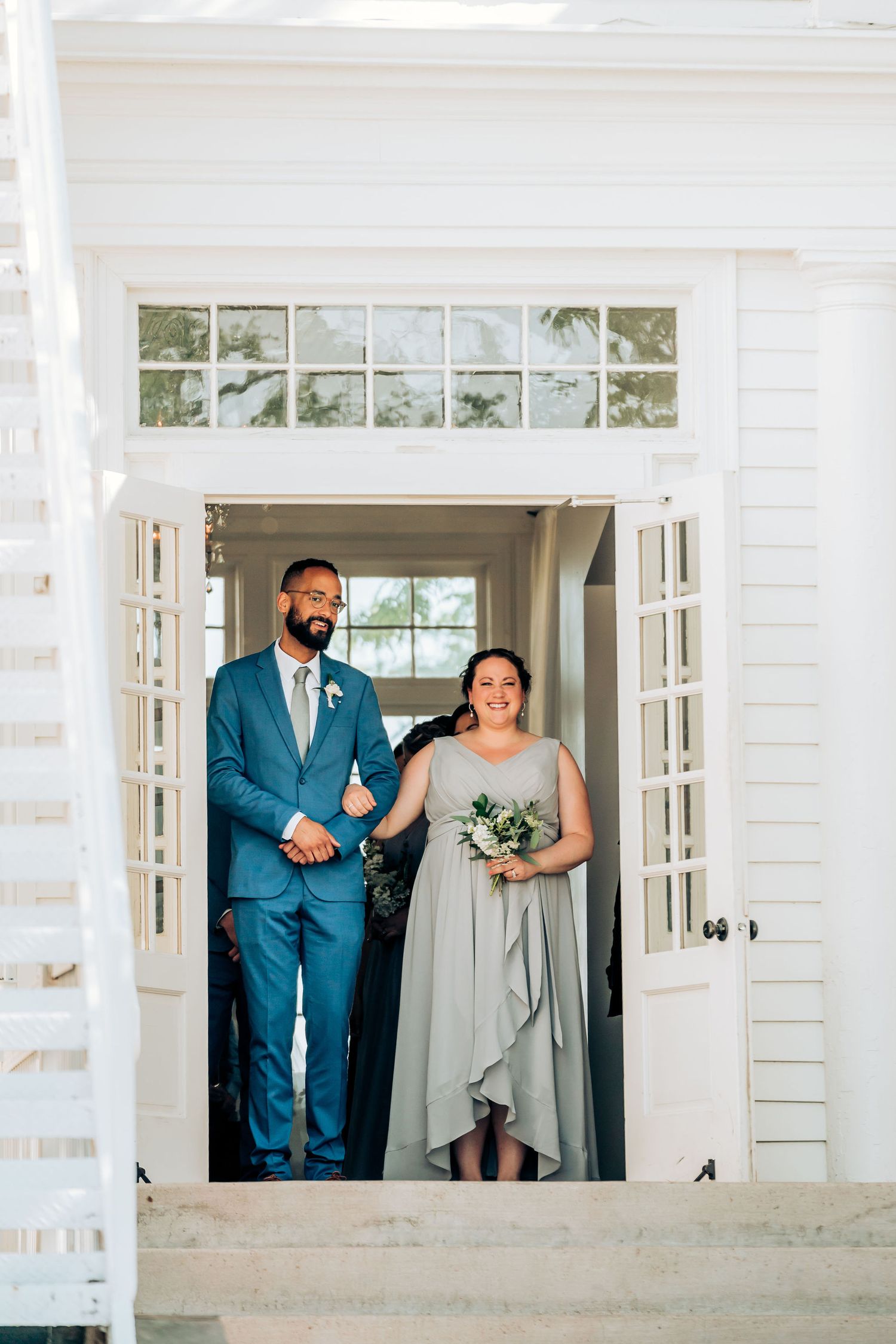 Wedding attendants pose in doorframe of white building, wearing blue and gray attire.