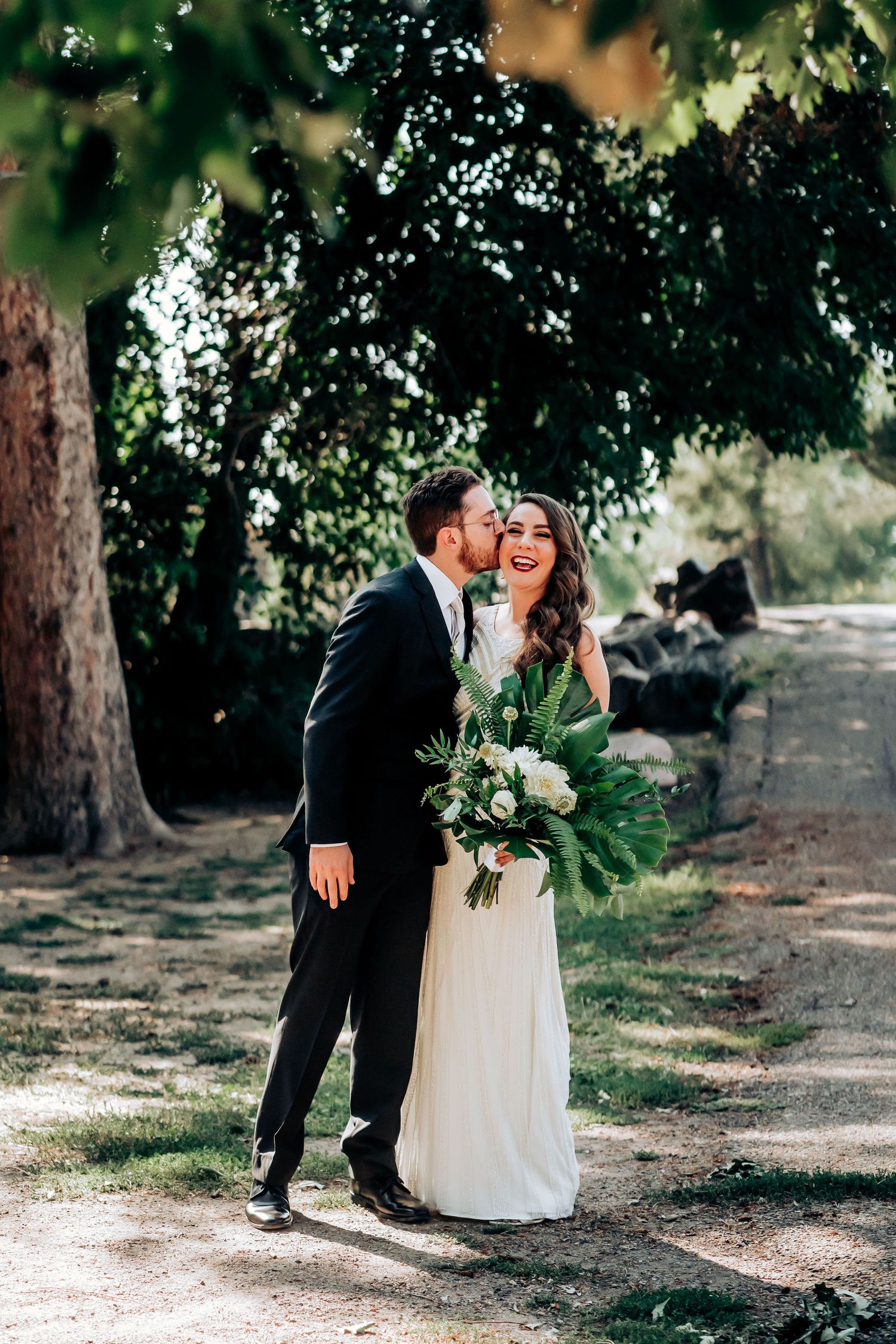 A romantic wedding moment captured under trees with the couple holding a lush green and white bouquet.