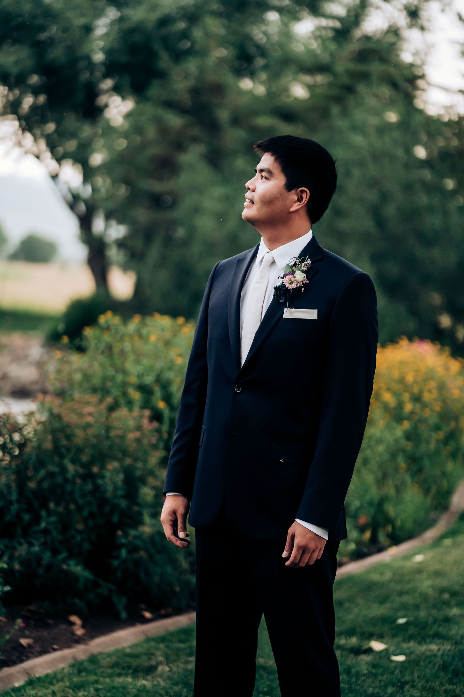 Groom in classic black tuxedo poses in garden with boutonniere.