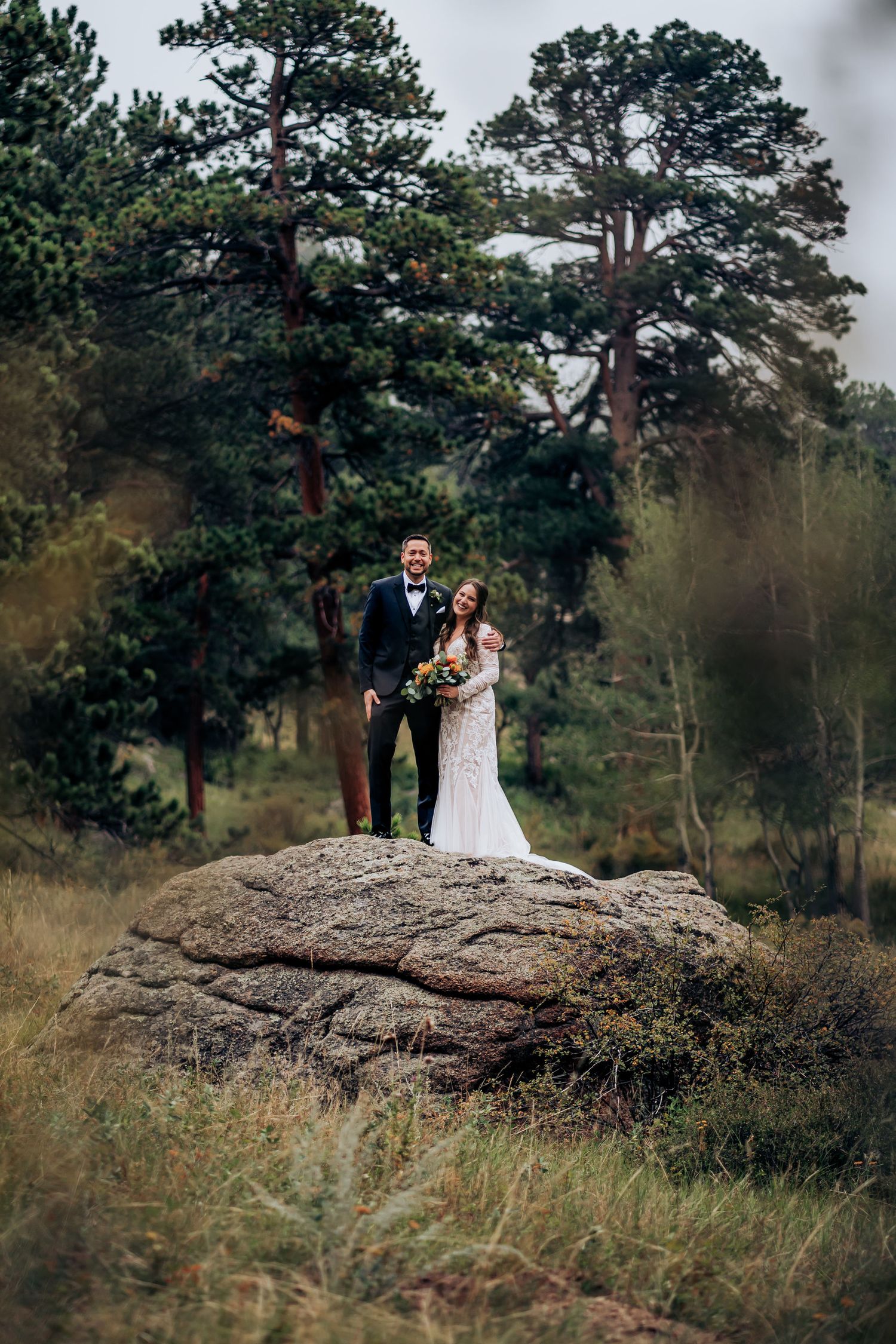 A wedding couple poses on a rocky outcrop with tall pine trees in the background.