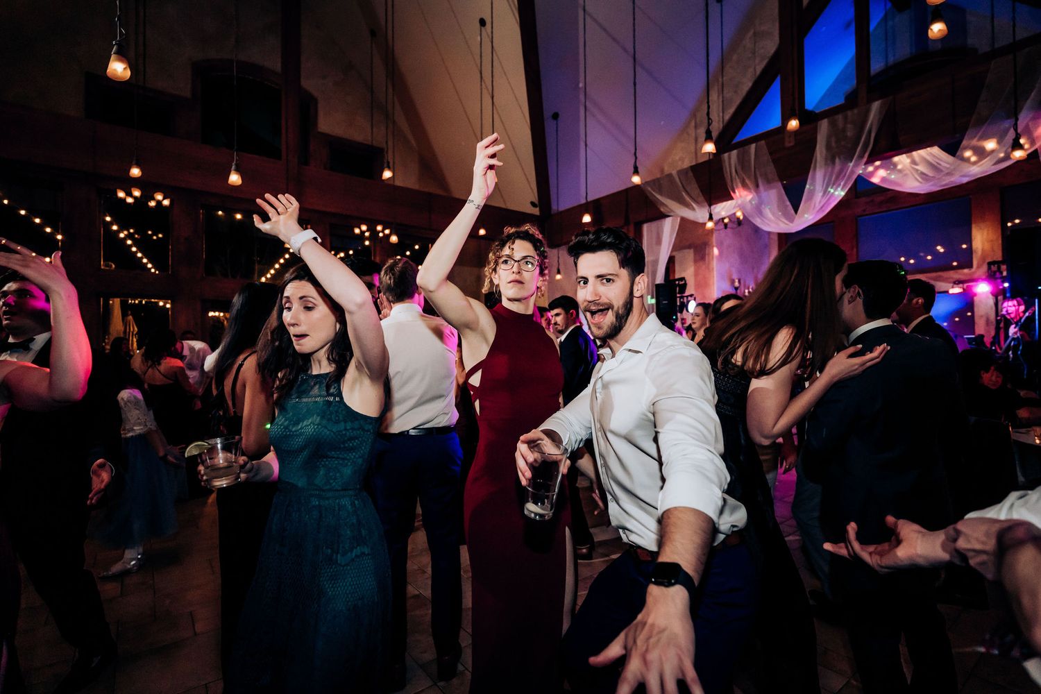 Energetic wedding guests dancing and celebrating with raised arms in a venue with mood lighting.