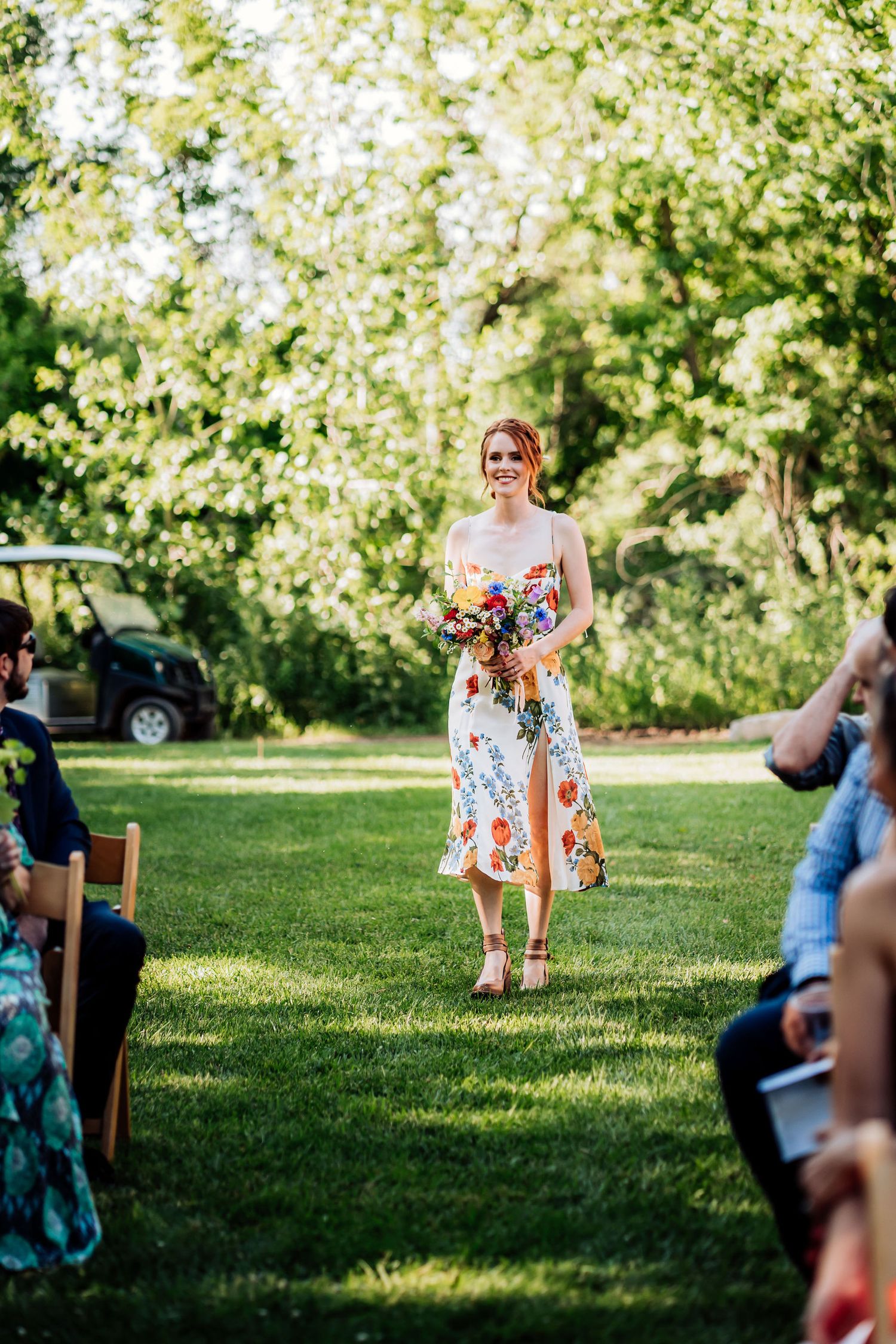 A guest in a floral summer dress walks down a grassy outdoor aisle at a garden wedding ceremony.