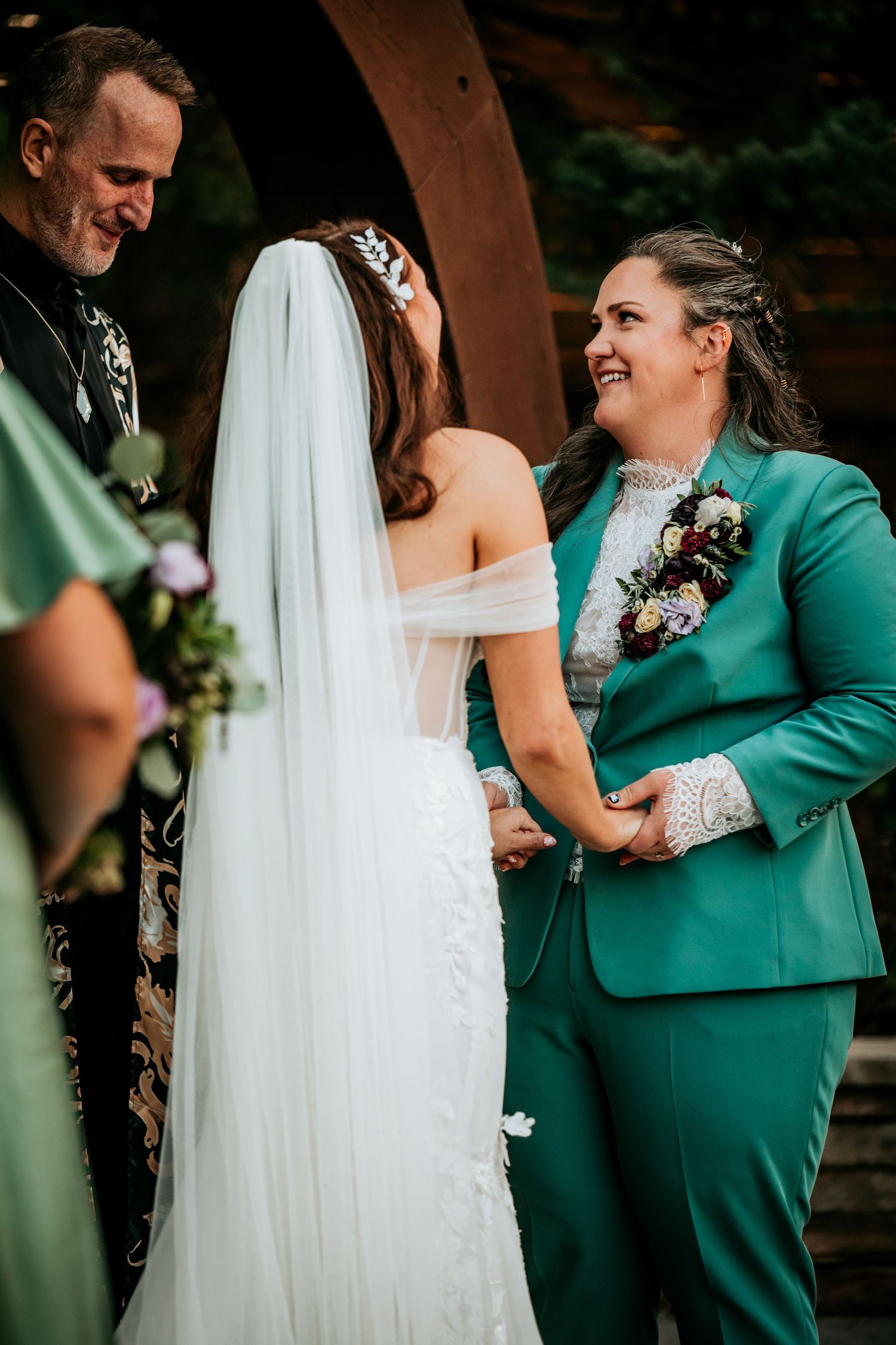 Bride in lace gown holds hands with partner in teal suit while exchanging vows at ceremony.
