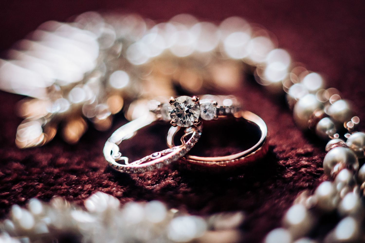 Diamond wedding rings and pearl necklace arranged on dark burgundy velvet surface with soft bokeh effect.