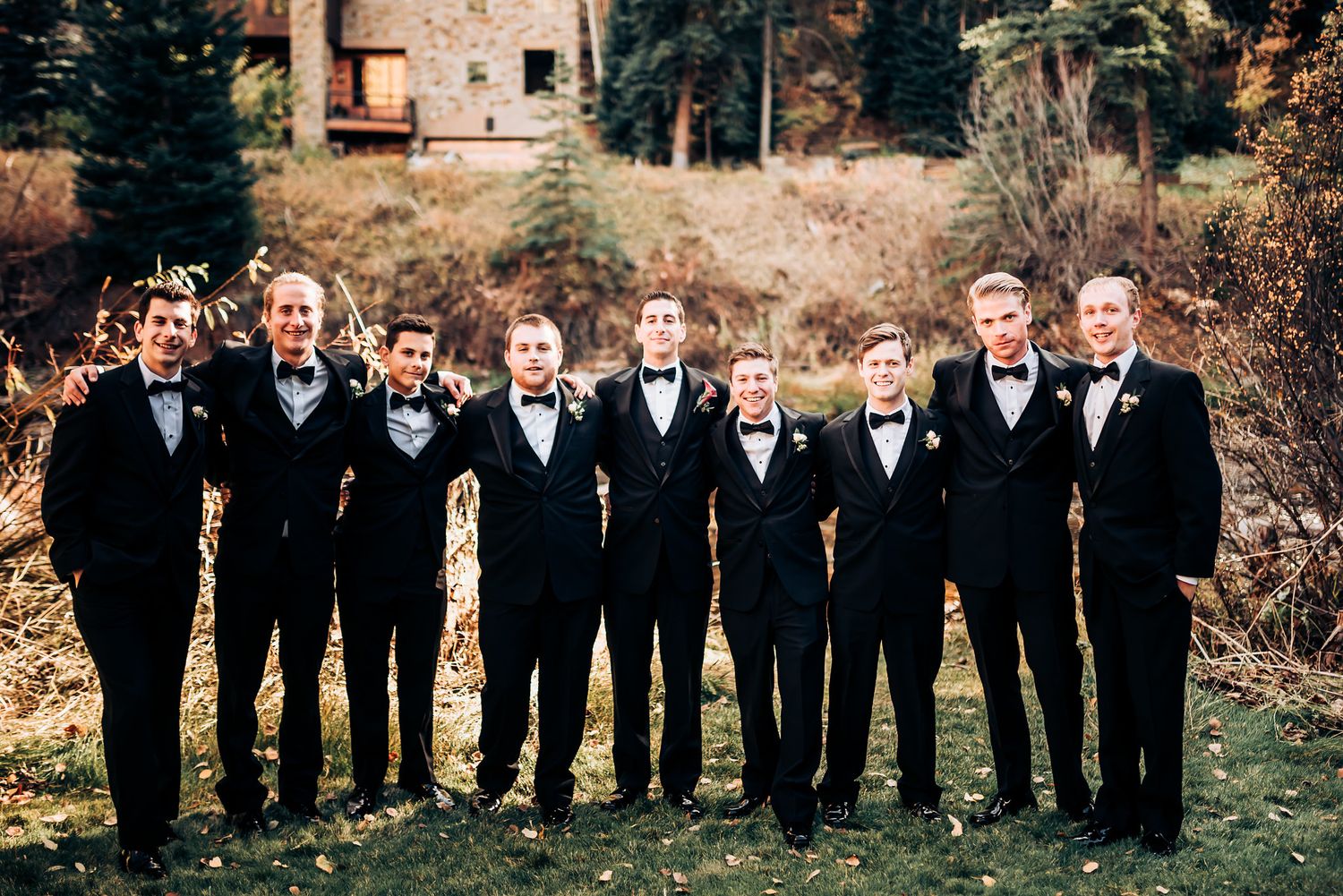 Row of groomsmen in black tuxedos stand on grass with rustic building backdrop.